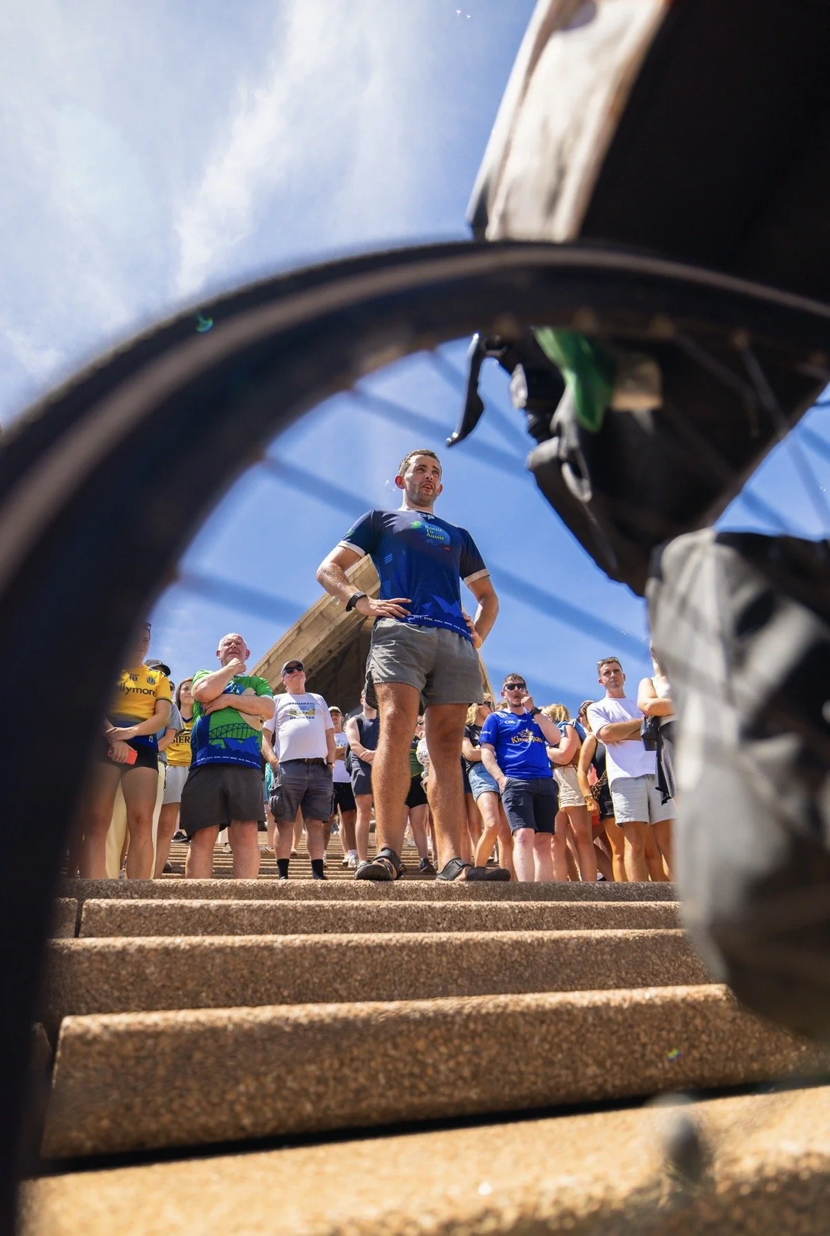 People gathered at the base of a large outdoor staircase, viewed through the opening of a bicycle wheel. The sky is blue with some clouds.