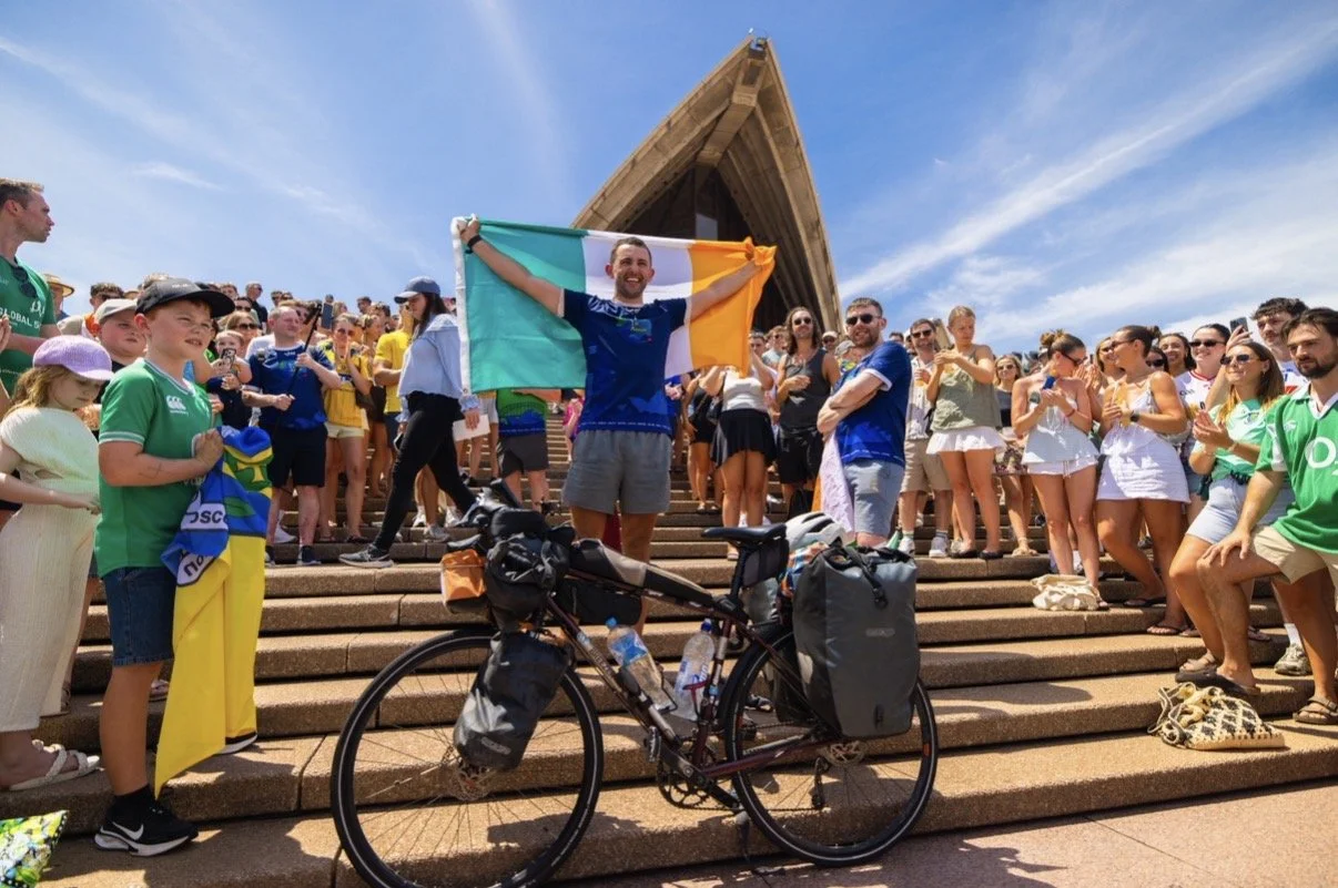 A young man holding an Irish flag stands at the top of stone steps in front of a building with a triangular roof, surrounded by a large crowd of people celebrating outdoors on a sunny day.