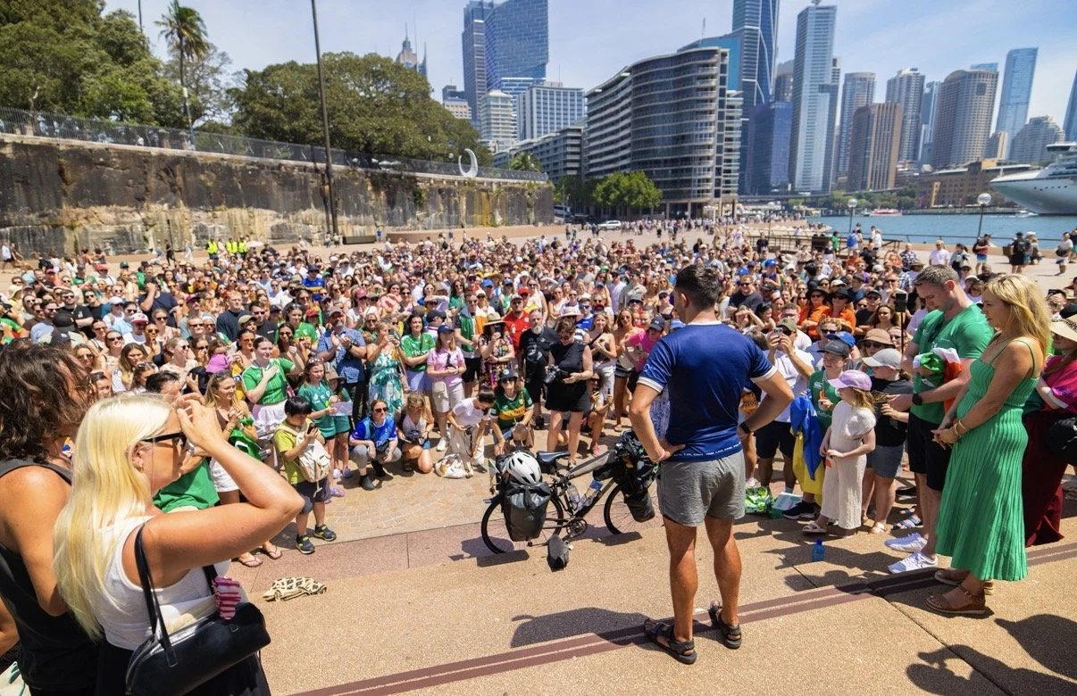A large crowd gathered outdoors in front of a city skyline with tall buildings, near water with boats, with some people taking photos and listening to a speaker, who is a man in a blue shirt standing with a bicycle.