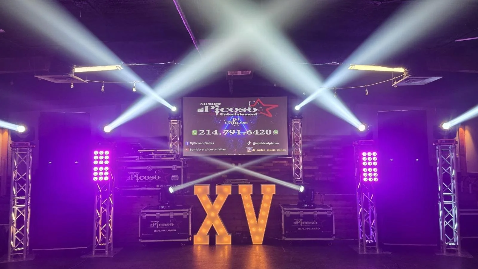 Stage setup with large illuminated Roman numerals XV, purple and white stage lighting, screens displaying entertainment info, and DJ equipment in the background.