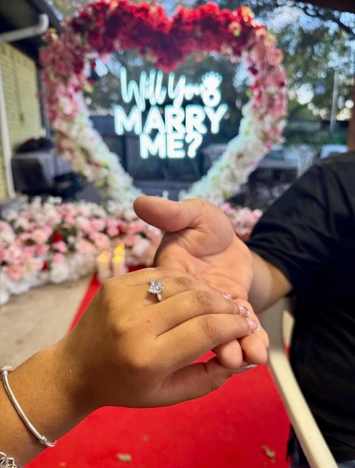 A close-up of a woman wearing a diamond engagement ring, holding hands with a man in front of a floral wedding arch and a neon sign that reads 'Will You Marry Me?'