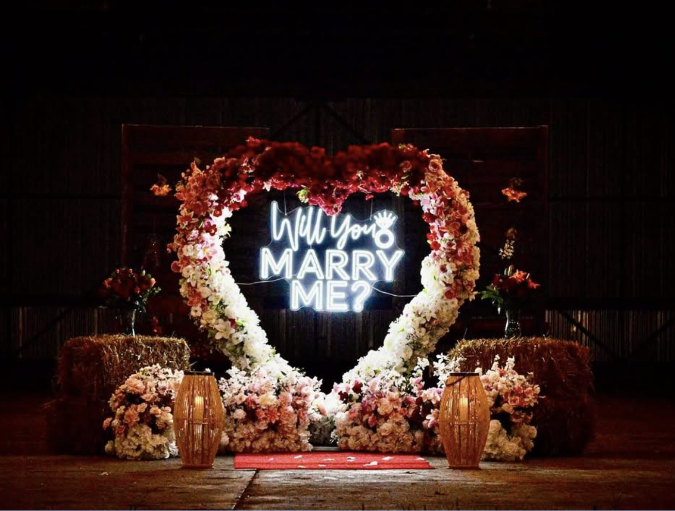 A decorative heart-shaped flower arrangement with a neon sign that reads 'Will You Marry Me?' in the center, flanked by vases and hay bales, staged for a marriage proposal.