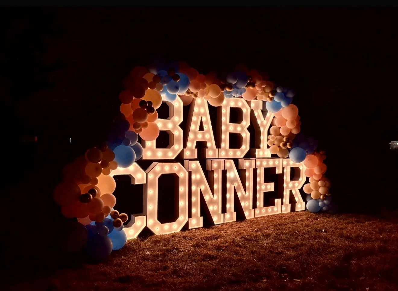 Illuminated sign that says "Baby Jonner" surrounded by colorful balloons, set against a dark background.