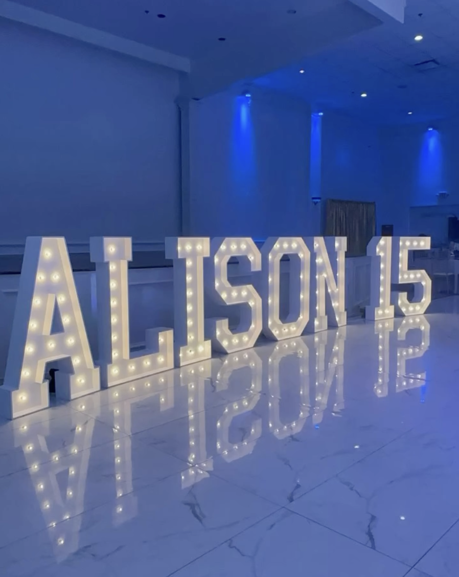 Large decorative marquee letters spelling out 'ALISON 15', illuminated with small light bulbs, placed on a polished floor in a dimly lit room with blue accent lighting.