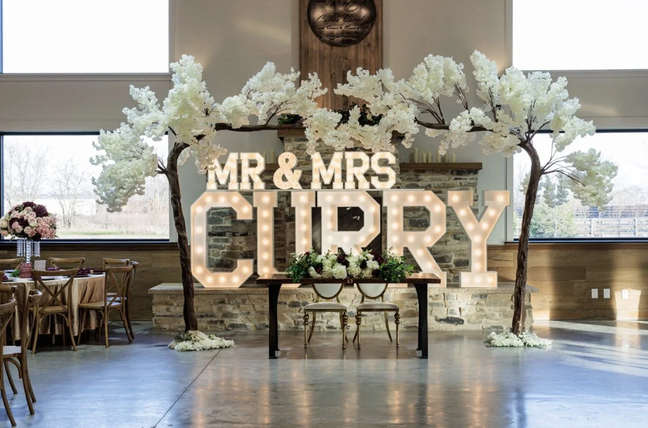 Decorative wedding setup with illuminated sign reading 'MR & MRS CURREY' flanked by artificial white trees with blossoms, and tables with flowers on the left side in a spacious room with large windows.