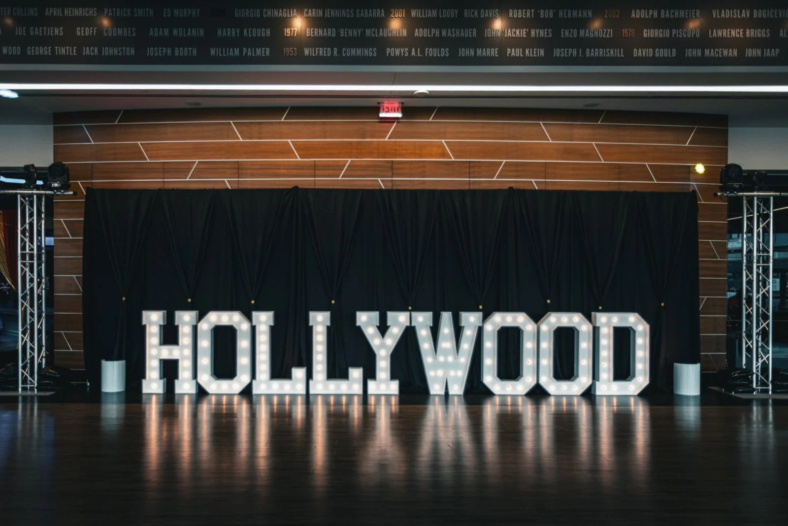 Large illuminated Hollywood sign decoration on a stage with black curtains, flanked by metal trusses, in a room with a wood-paneled wall. A list of names and years is displayed on a panel above.