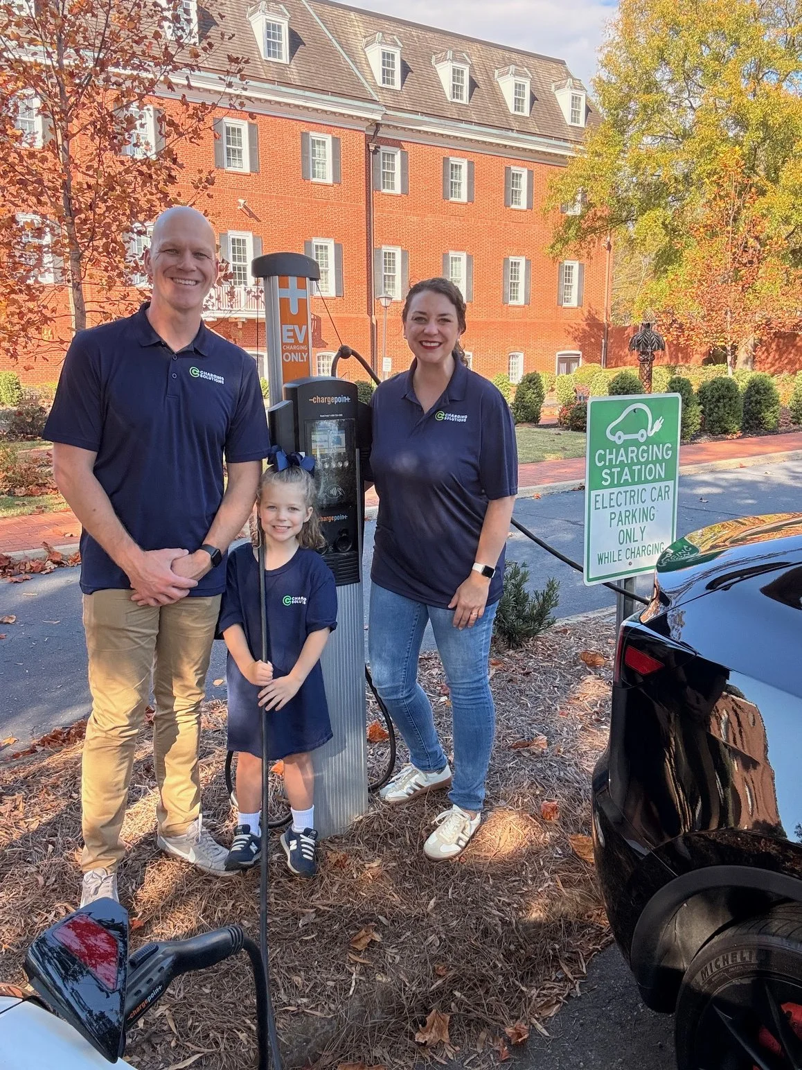 Business owners and their daughter stand next to an electric vehicle charging station in a parking lot with an orange brick building and trees with autumn leaves in the background. The group appears happy and the scene is set during daytime.