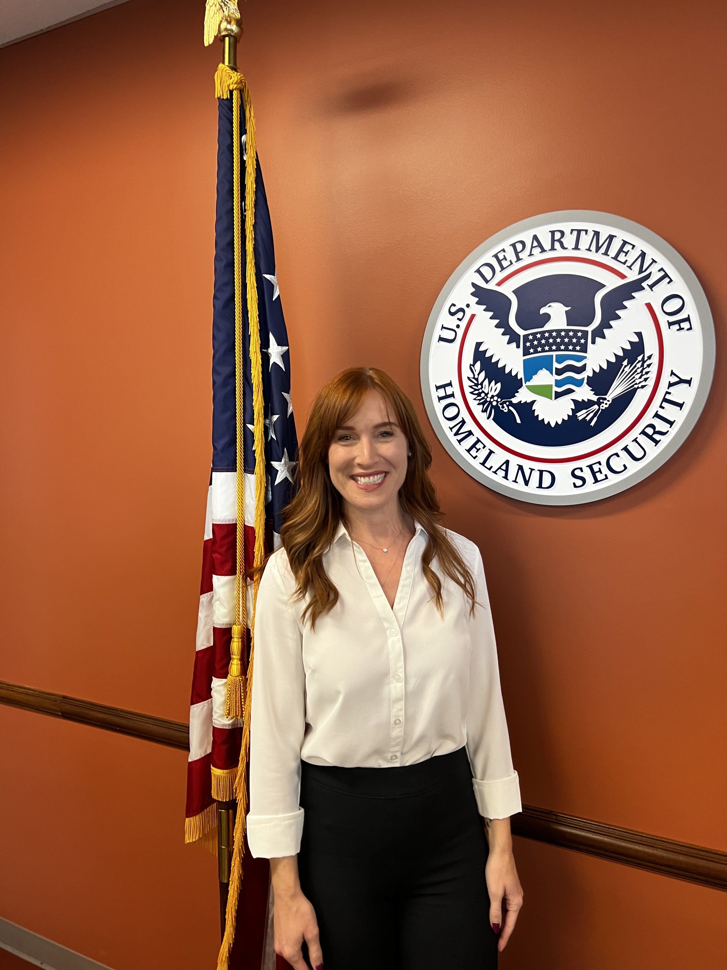 A woman stands smiling in front of a U.S. flag and the Department of Homeland Security seal