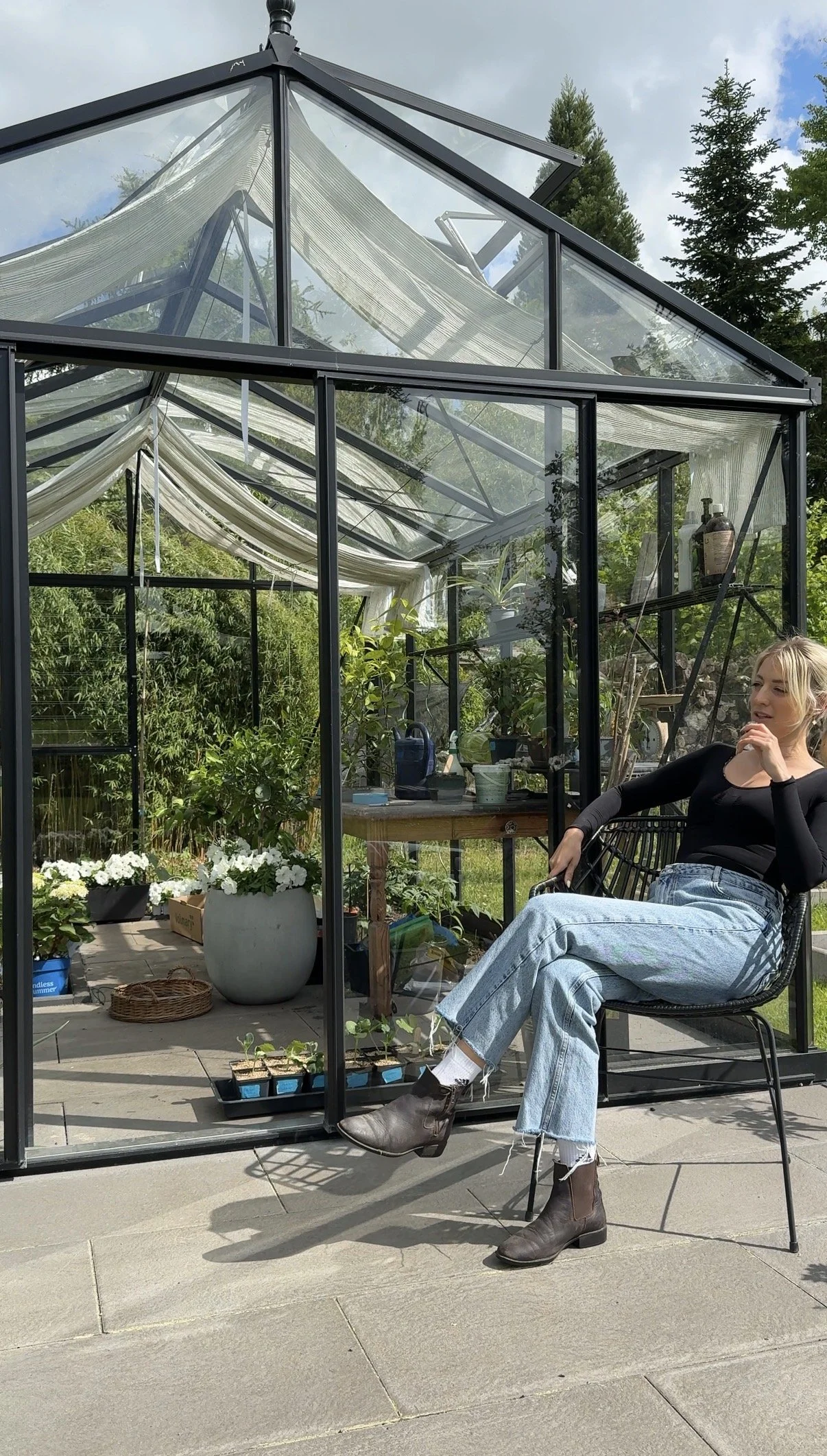 Garten Creatorin Janet Hirse von Hannis_Haus sitzt vor ihrem Gewächshaus zwischen Pflanzen und filmt authentischen Gartencontent.