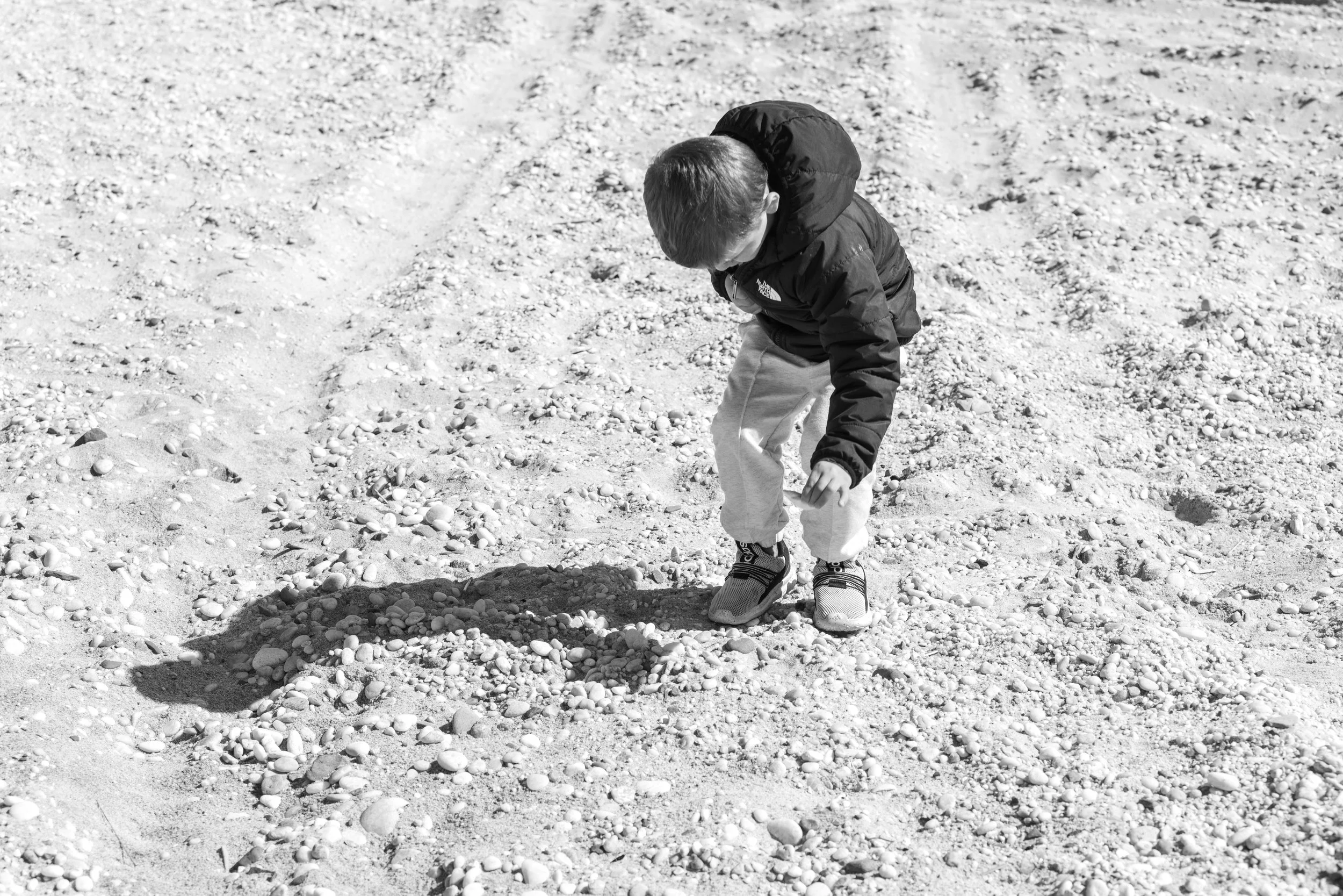 A young boy wearing a black jacket, light-colored pants, and sneakers is crouched down on a gravelly outdoor surface, looking at or picking up small stones.