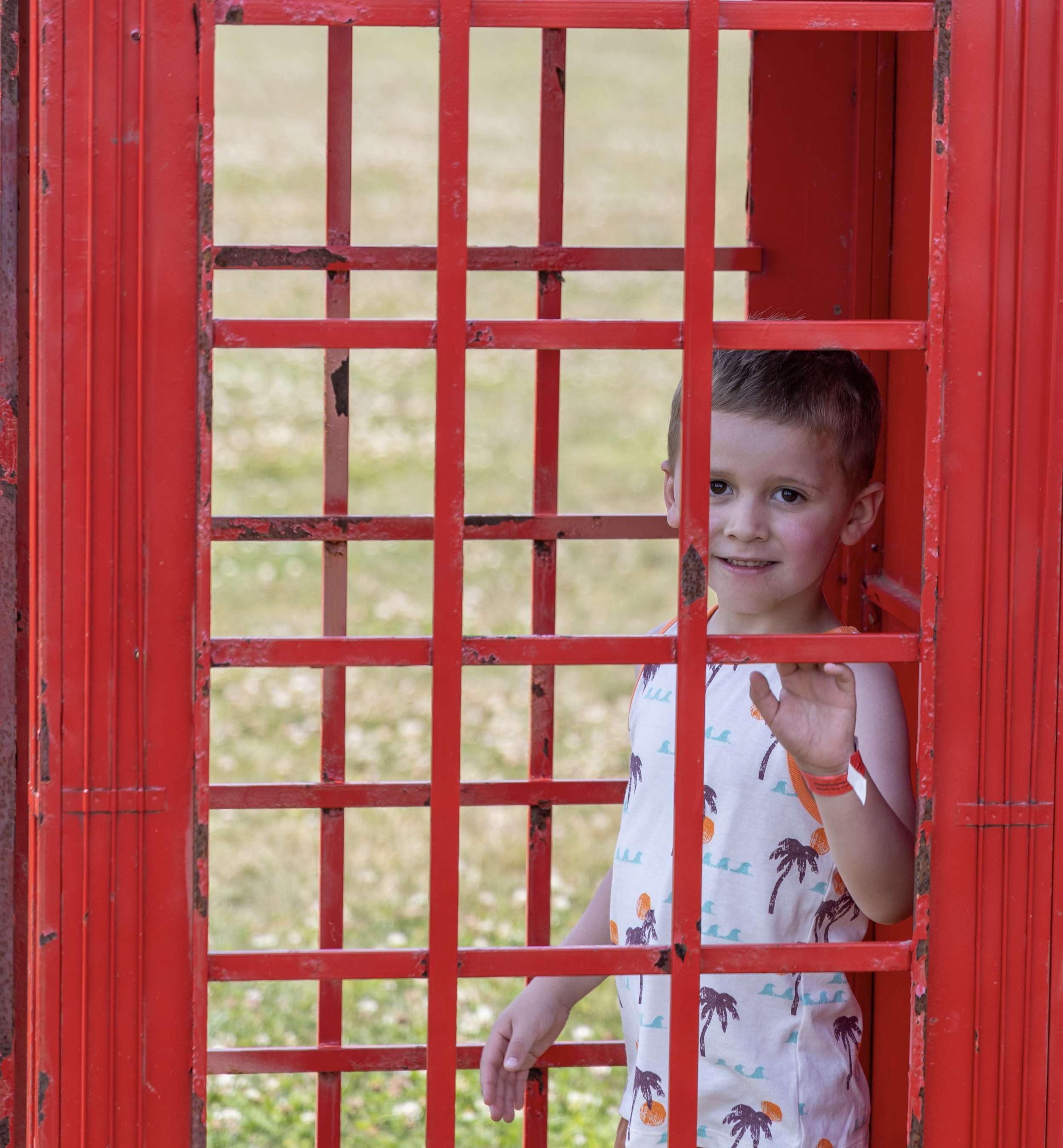 A young boy with light brown hair and dark eyes peeking through a red metal gate with vertical and horizontal bars. The boy is wearing a white sleeveless shirt with palm tree and pineapple patterns, and he has a small wristband on his left wrist. The background is blurred with grass and trees.