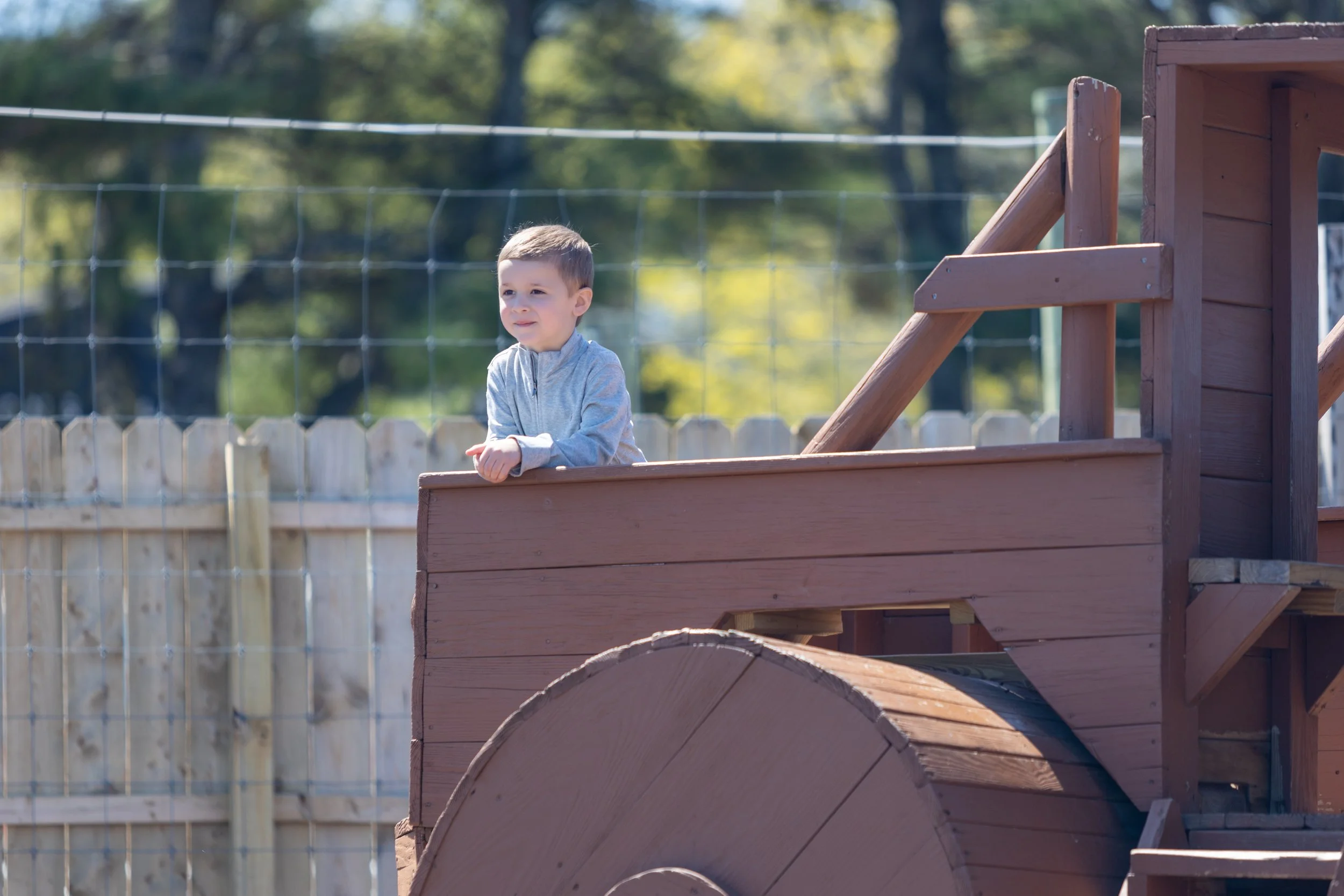 A young boy in a gray hoodie standing on a wooden playground structure with steps and a slide, outdoors with a fence and trees in the background.