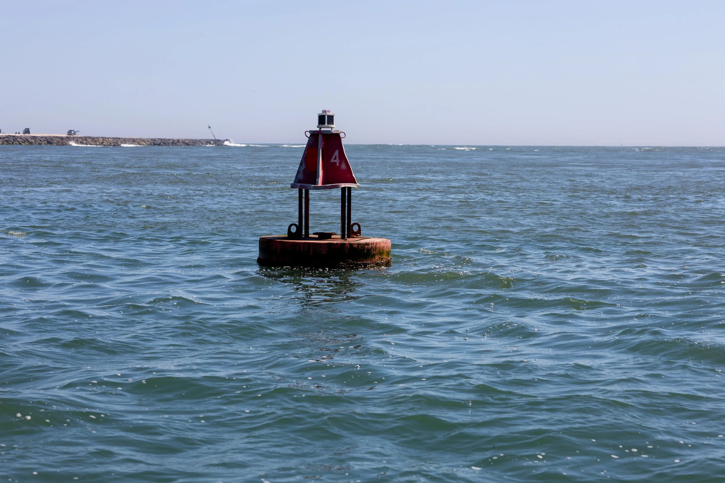 A red buoy with the number 4 on it floating in a body of water near a rocky shoreline under a clear blue sky.