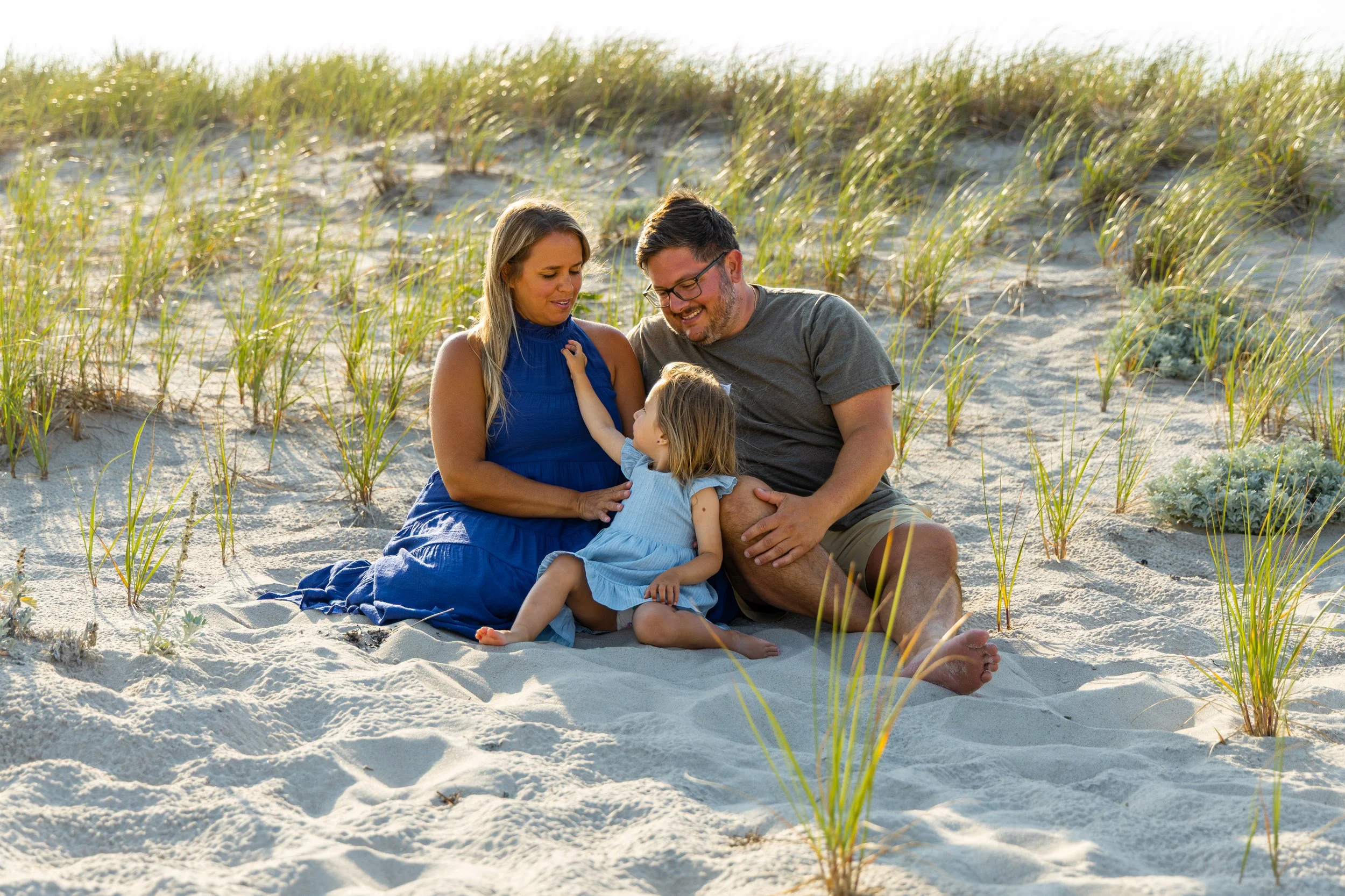 A family of three sitting on the beach surrounded by grass, enjoying a sunny day.