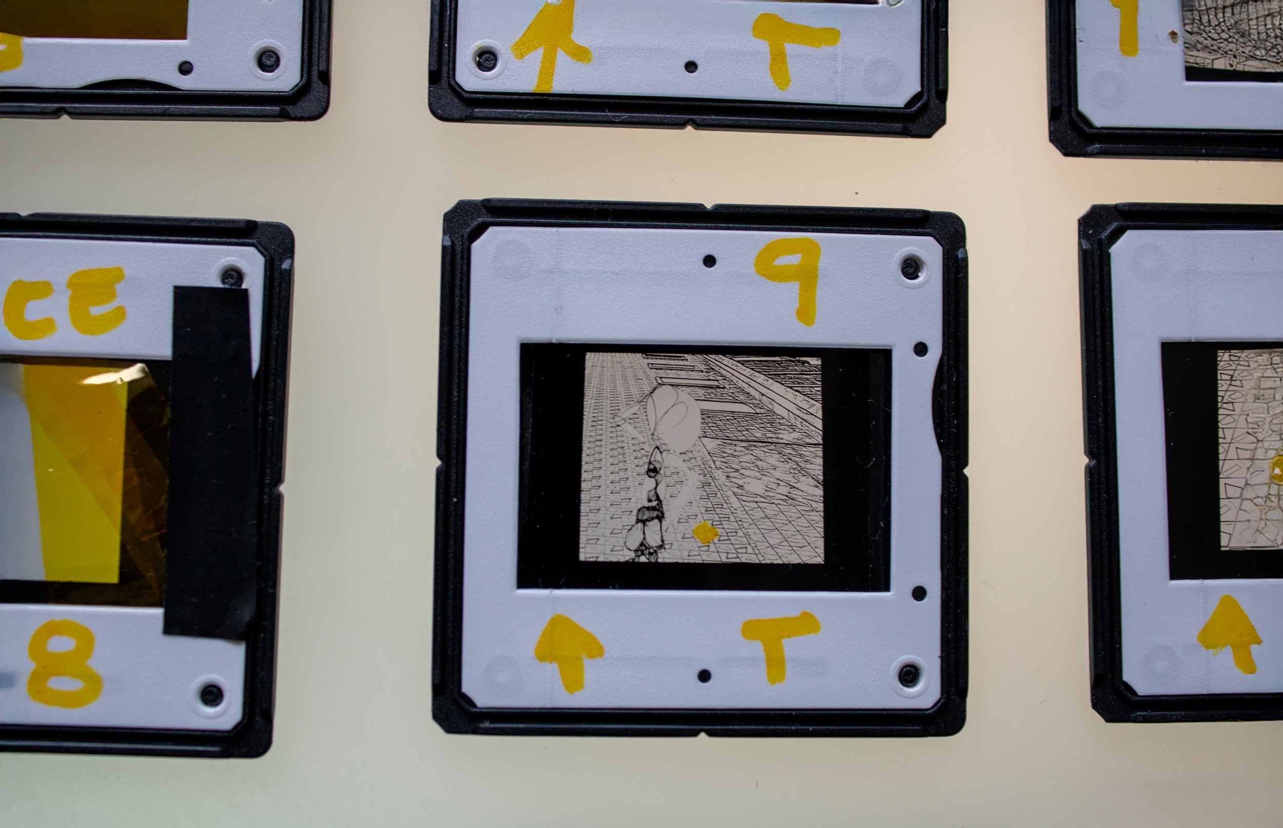 Slide fotográfico analógico em preto e branco da série Slides – Fotografia, de Sandra Unglert Fernandes, mostrando figura humana diante de edifício e pequeno círculo amarelo.
