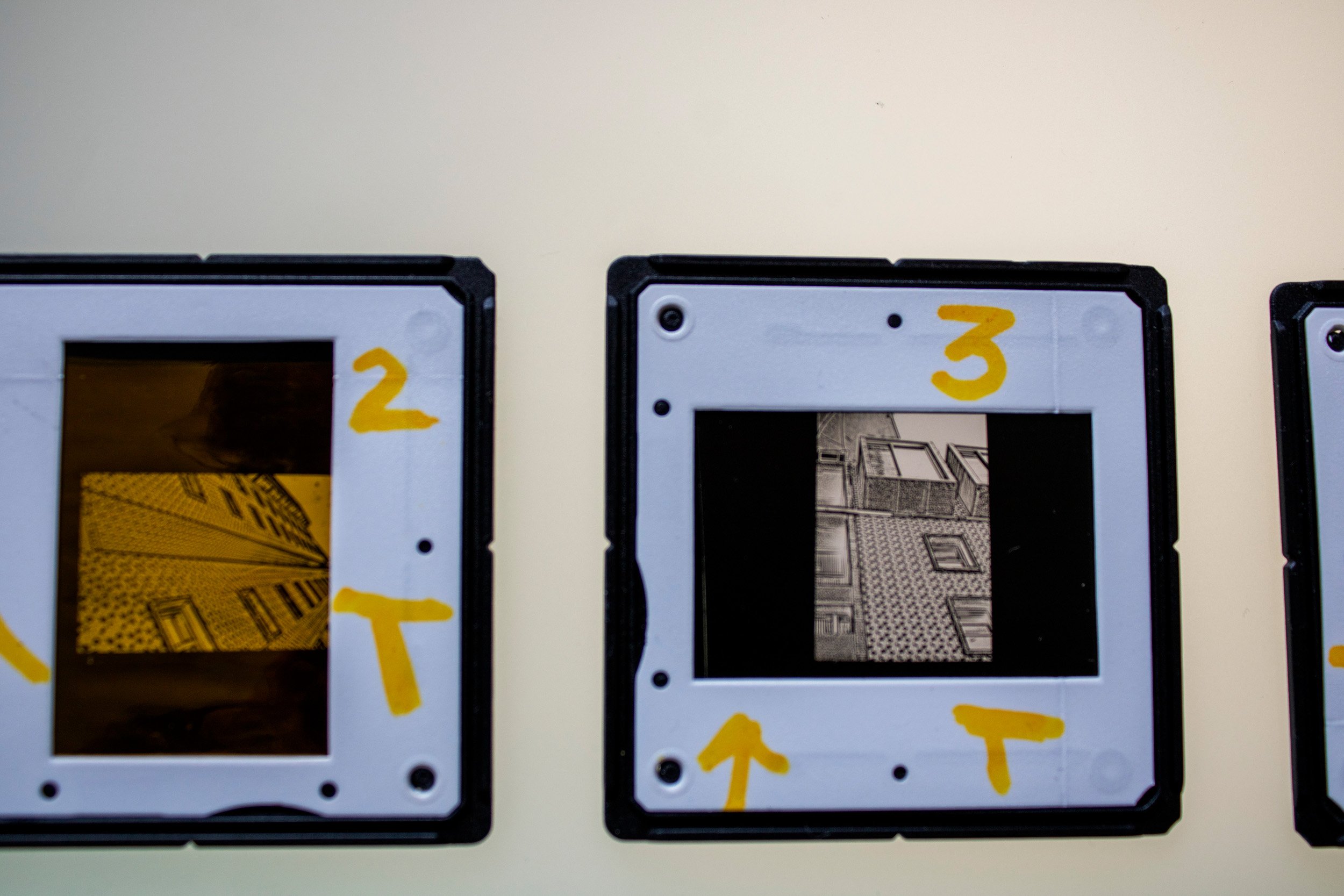 Dois slides fotográficos analógicos em preto e branco, integrantes da série Slides – Fotografia, de Sandra Unglert Fernandes, mostrando detalhes de edifícios, um deles com intervenção em papel celofane amarelo e ambos com marcações manuscritas.