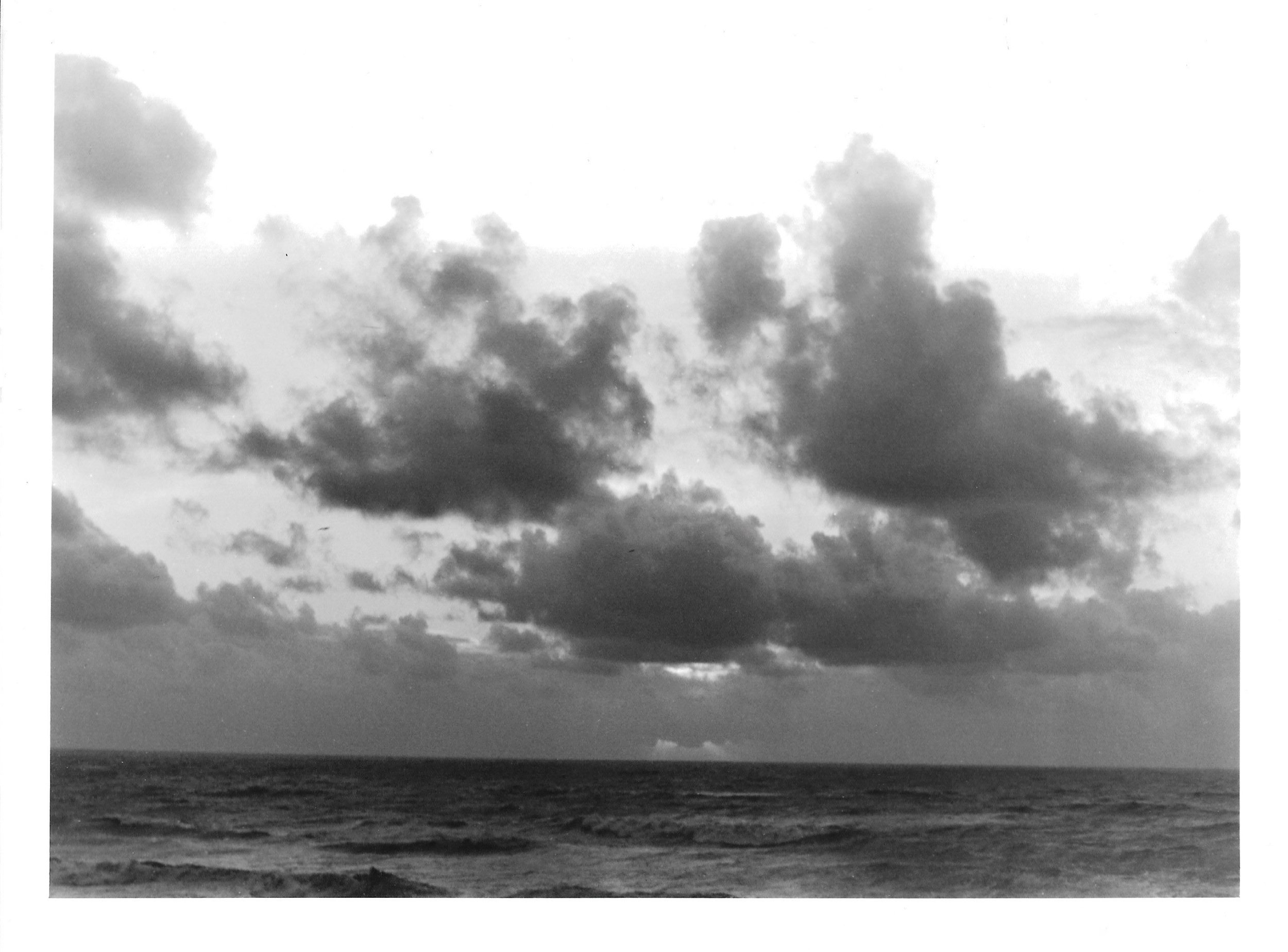 Fotografia analógica em preto e branco, integrante da Série 2, de Sandra Unglert Fernandes, mostrando o mar sob um céu com nuvens volumosas e formações atmosféricas contrastantes.