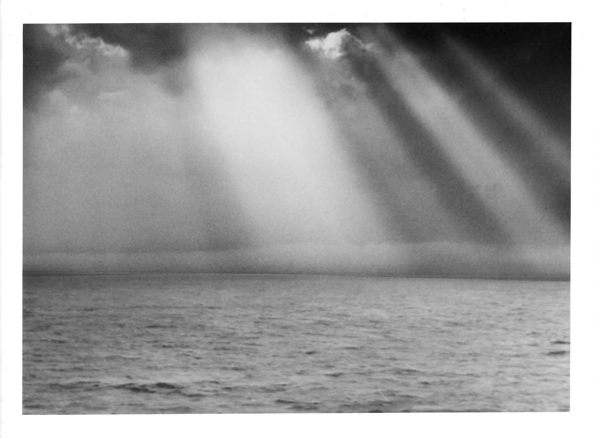Fotografia analógica em preto e branco, integrante da Série 2, de Sandra Unglert Fernandes, mostrando feixes de luz atravessando as nuvens e iluminando o mar.