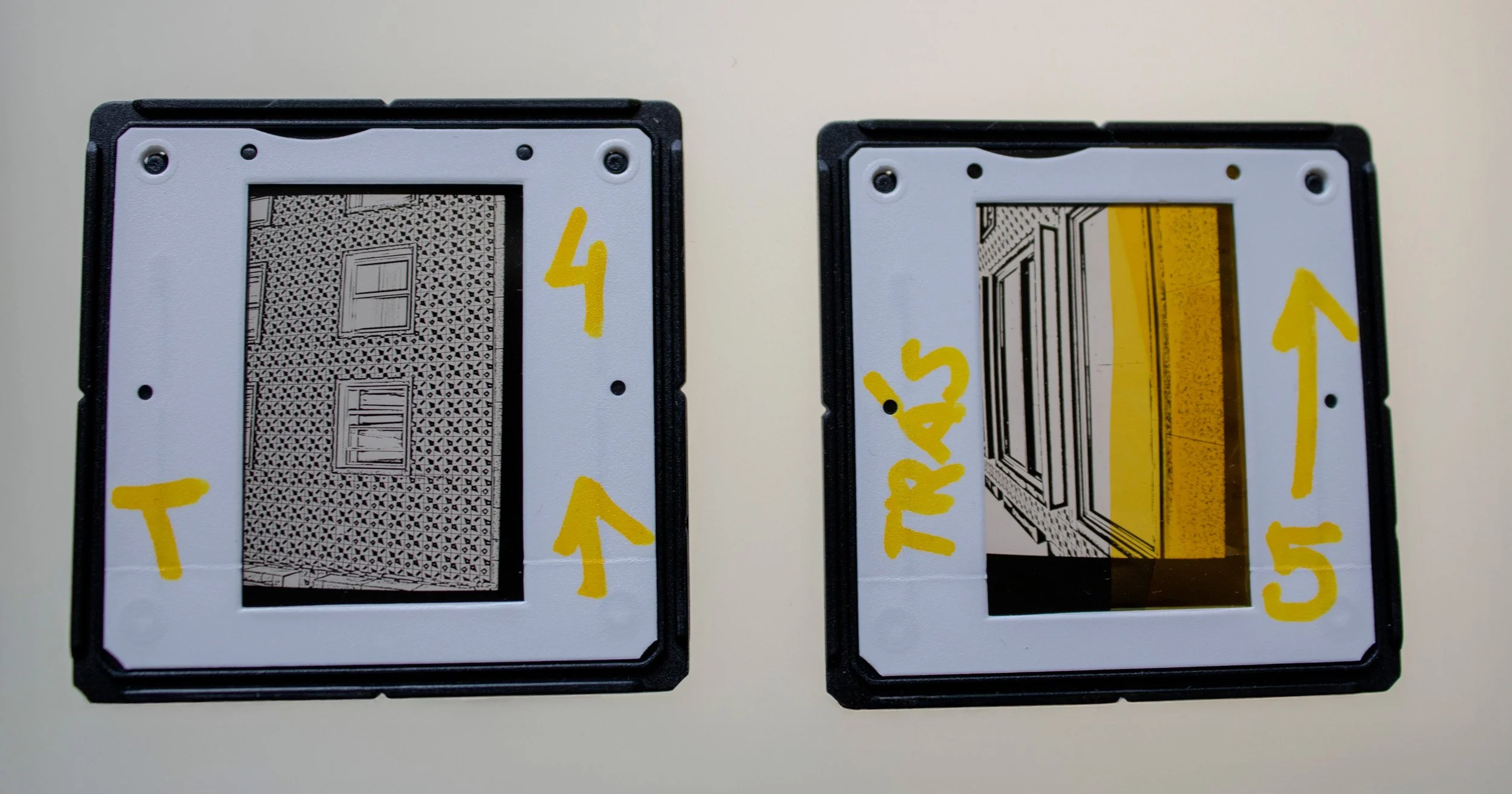 Dois slides fotográficos analógicos em preto e branco, integrantes da série Slides – Fotografia, de Sandra Unglert Fernandes, mostrando detalhes de edifícios, um deles com intervenção em papel celofane amarelo e ambos com marcações manuscritas.