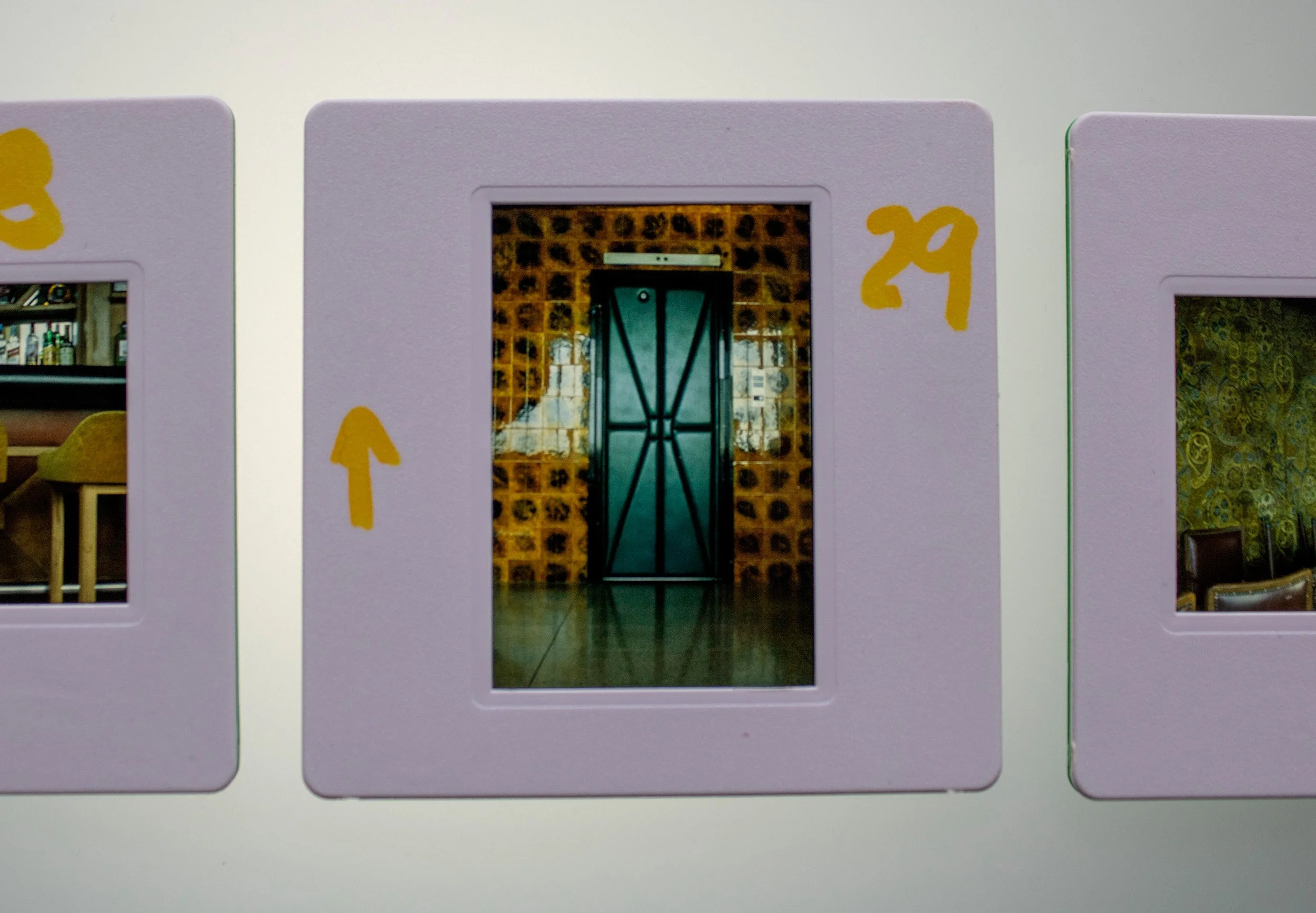 Slide fotográfico analógico colorido, integrante da série Slides – Fotografia, de Sandra Unglert Fernandes, mostrando o interior de um café com bancos, intervenções em papel celofane amarelo e marcações manuscritas.