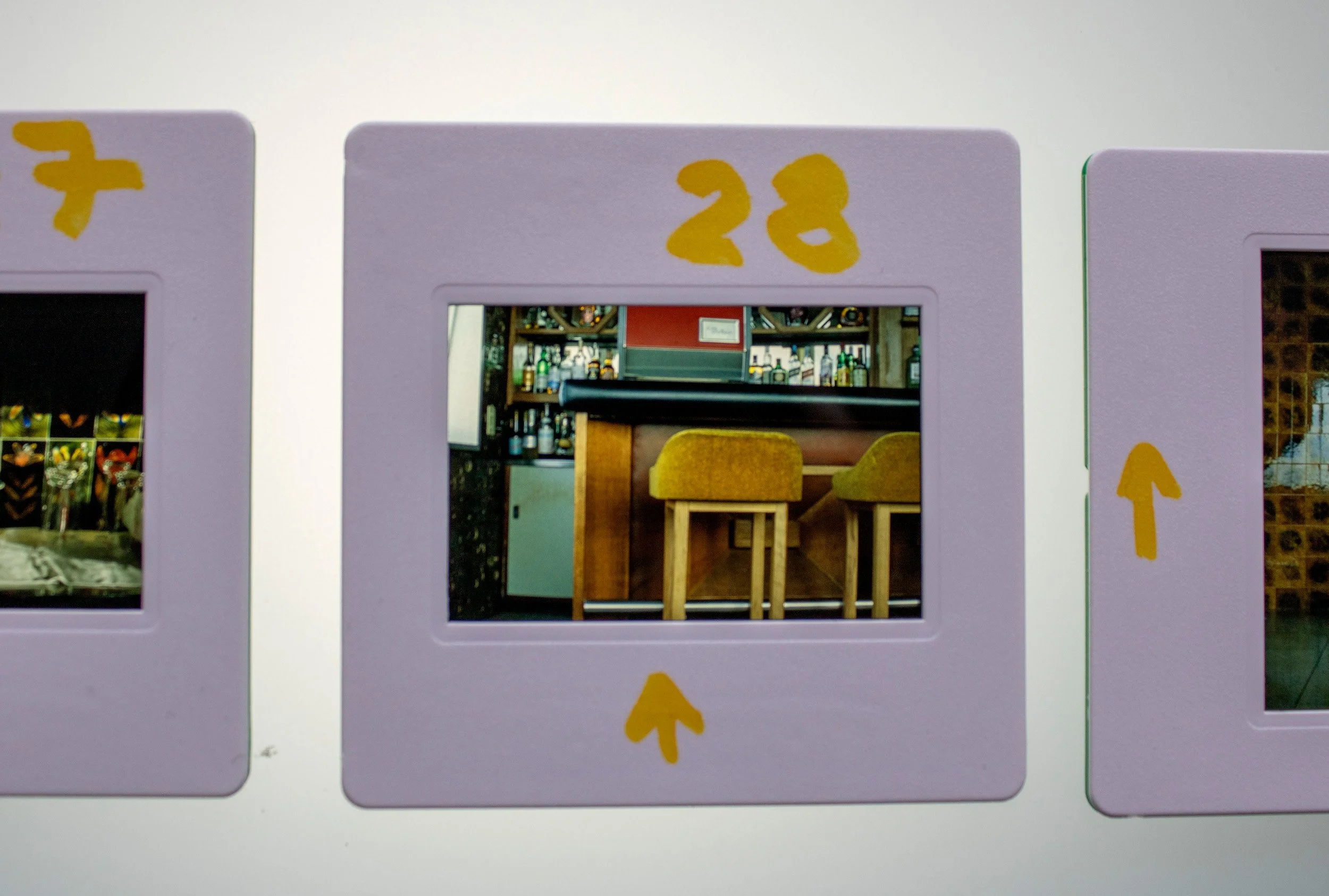 Slide fotográfico analógico colorido, integrante da série Slides – Fotografia, de Sandra Unglert Fernandes, mostrando o interior de um café com bancos, intervenções em papel celofane amarelo e marcações manuscritas.