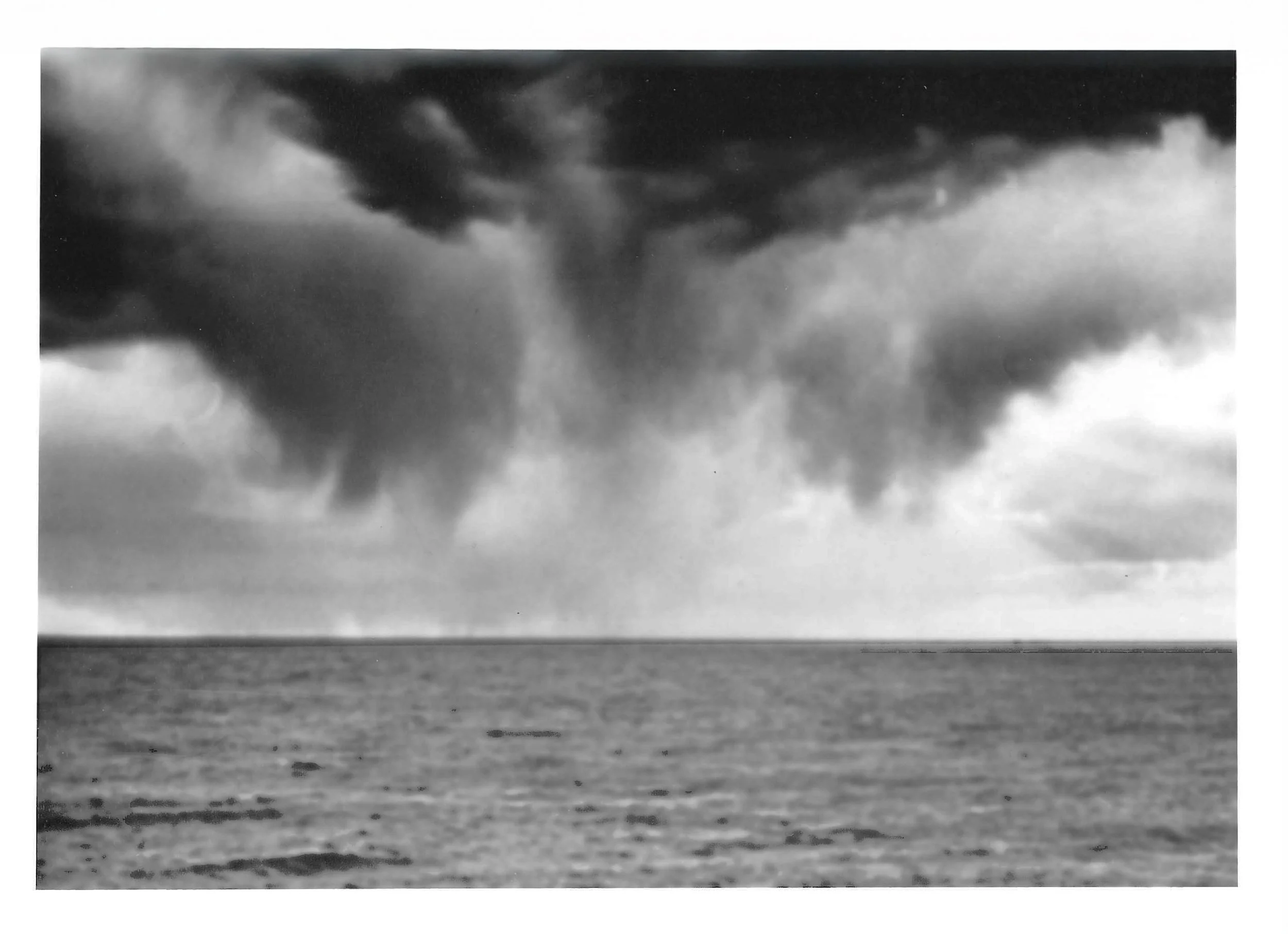 Fotografia analógica em preto e branco, integrante da Série 2, de Sandra Unglert Fernandes, mostrando nuvens densas sobre o mar e uma cortina de chuva no horizonte.