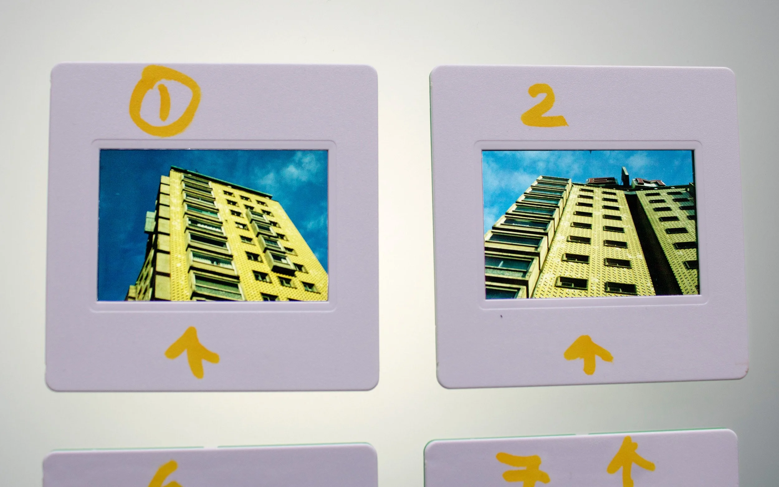 Slides fotográficos analógicos coloridos, integrantes da série Slides – Fotografia, de Sandra Unglert Fernandes, mostrando imagens de edifícios em perspectiva com intervenções em papel celofane amarelo e marcações manuscritas.