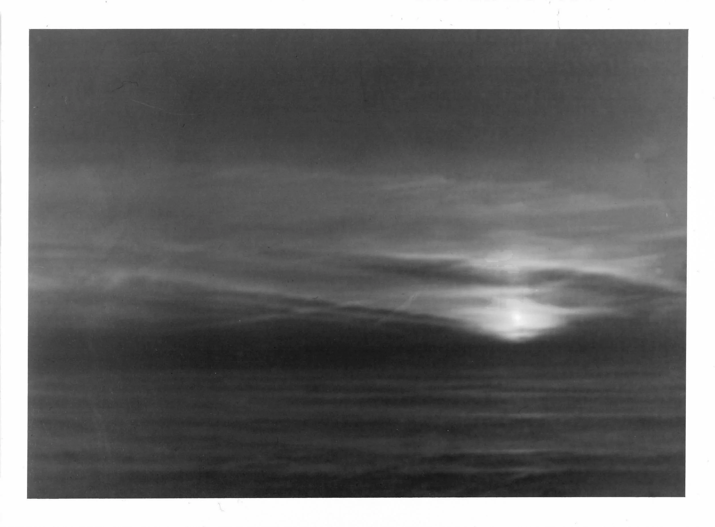 Fotografia analógica em preto e branco da Série 1, de Sandra Unglert Fernandes, mostrando o sol parcialmente encoberto por nuvens sobre o mar.