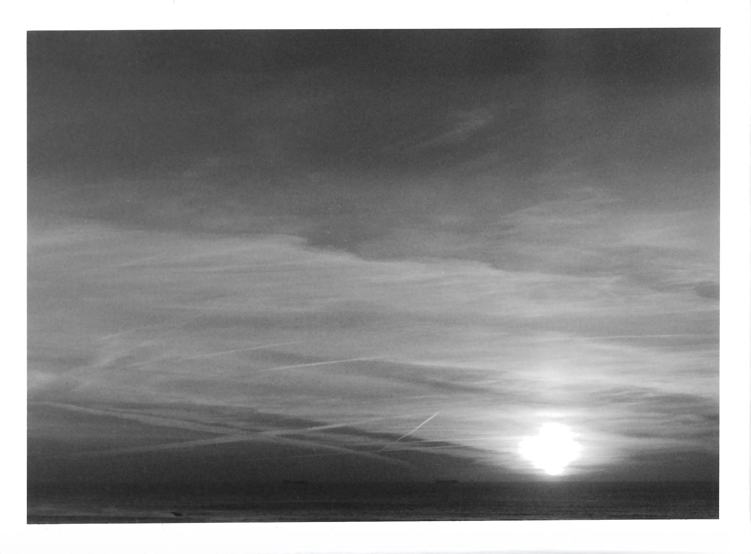 Fotografia analógica em preto e branco, integrante da Série 1, de Sandra Unglert Fernandes, mostrando o sol sobre o mar, com reflexo na água, nuvens e rastros lineares no céu.