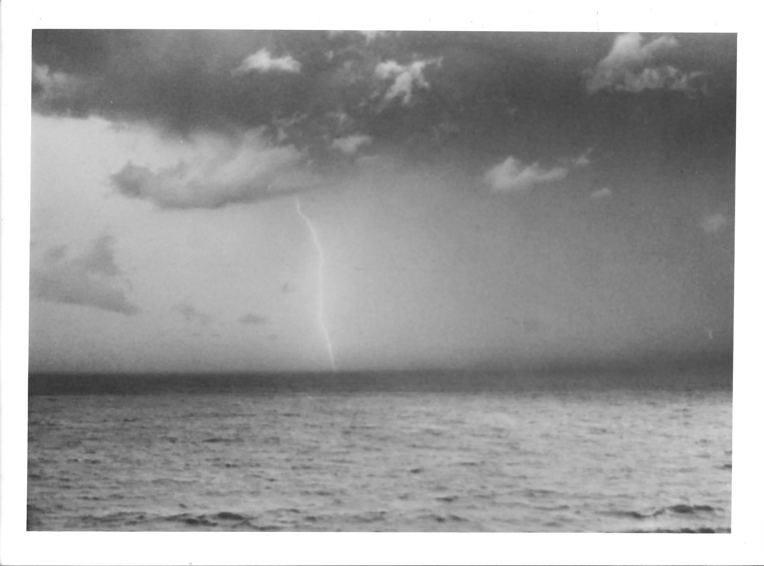 Fotografia analógica em preto e branco, integrante da Série 2, de Sandra Unglert Fernandes, mostrando um raio sobre o mar sob céu carregado de nuvens.