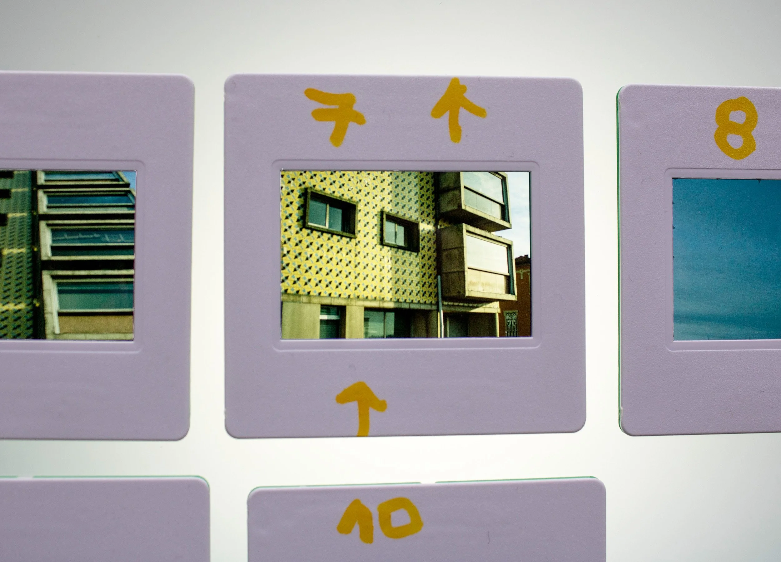 Slide fotográfico analógico coloridos, integrante da série Slides – Fotografia, de Sandra Unglert Fernandes, mostrando a fachada de um edifício, com intervenções em papel celofane amarelo e marcações manuscritas.