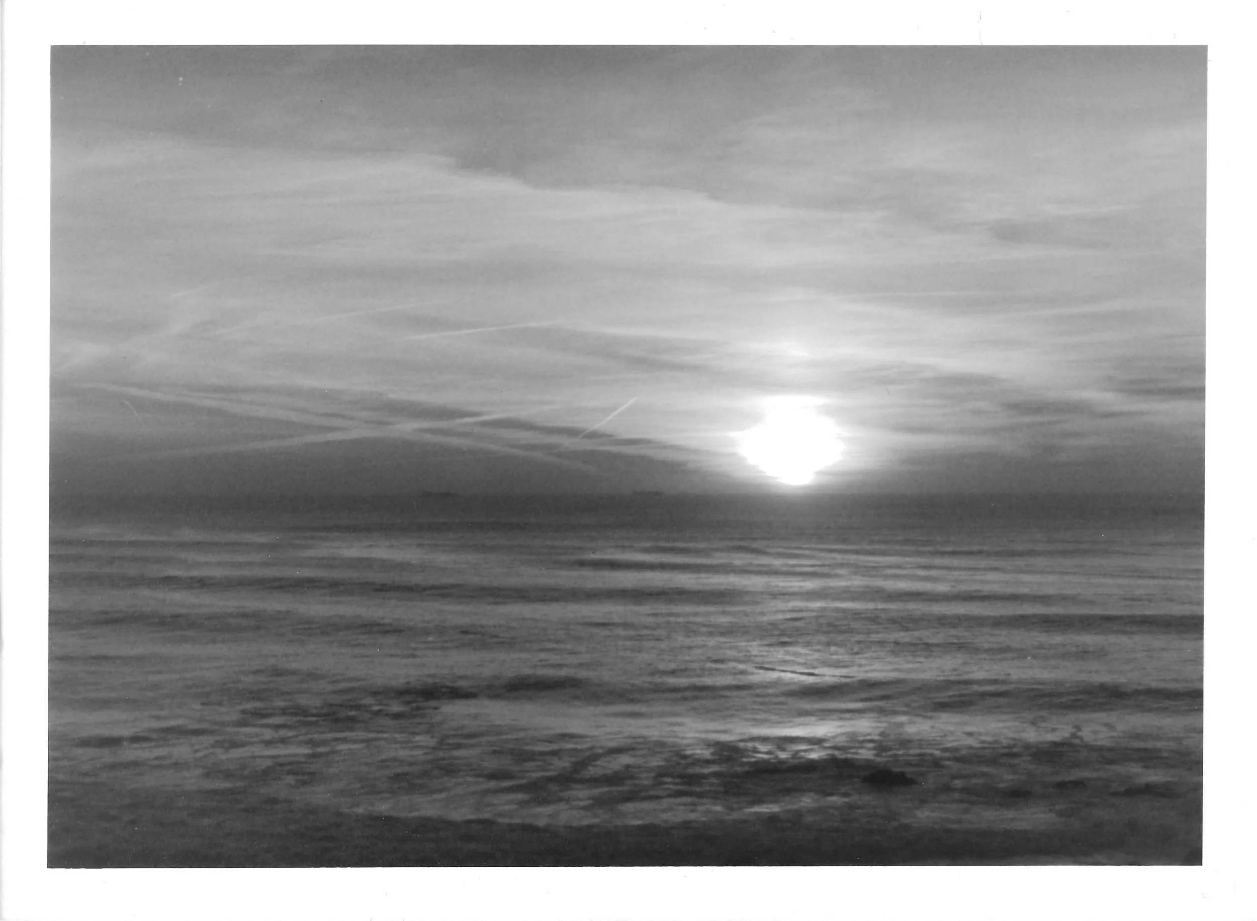 Fotografia analógica em preto e branco da Série 1, de Sandra Unglert Fernandes, mostrando o sol sobre o mar, com reflexo na água, nuvens e rastros lineares no céu.