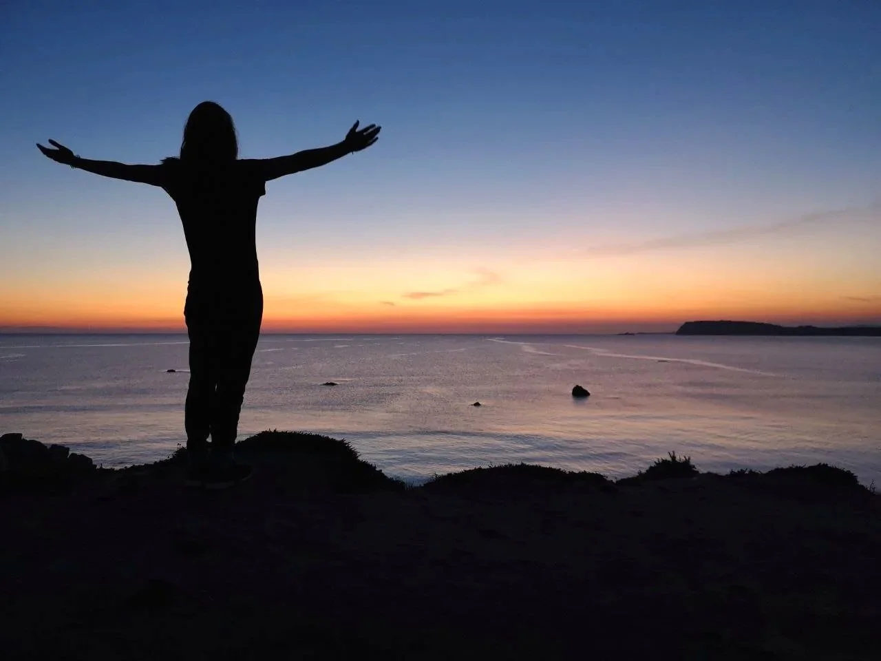 Eine silhouettierte Person mit ausgebreiteten Armen steht an der Küste während des Sonnenuntergangs, Blick auf das Meer und den Himmel mit orange-rosa Farbverläufen.