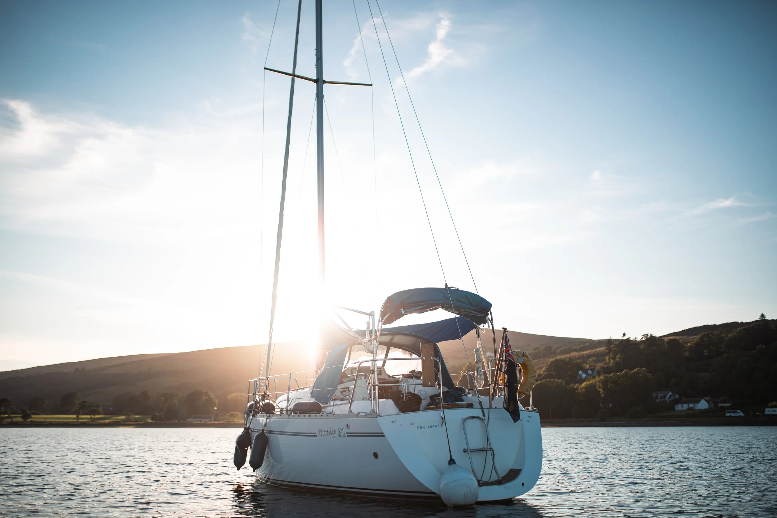 A white sailboat floating on calm water with a hill and houses in the background during sunset or sunrise.
