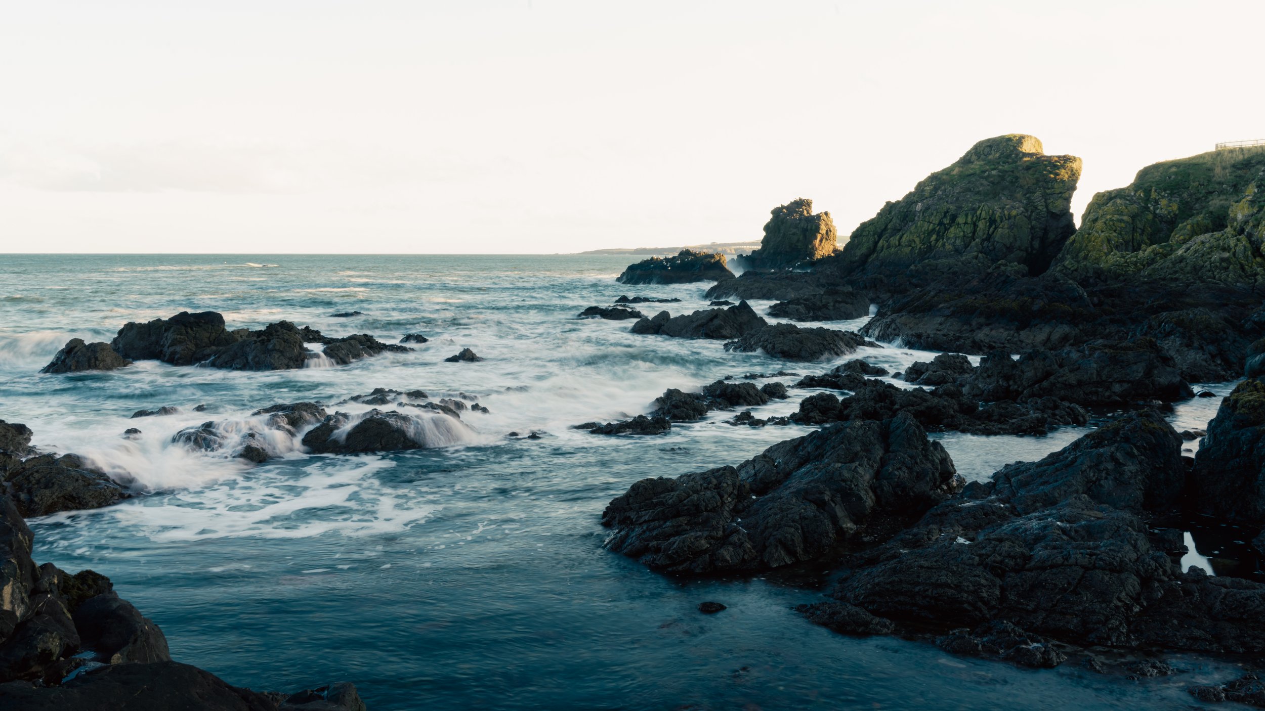 Coastal scene with rocky shoreline, waves crashing against rocks, and large moss-covered cliffs in the distance.
