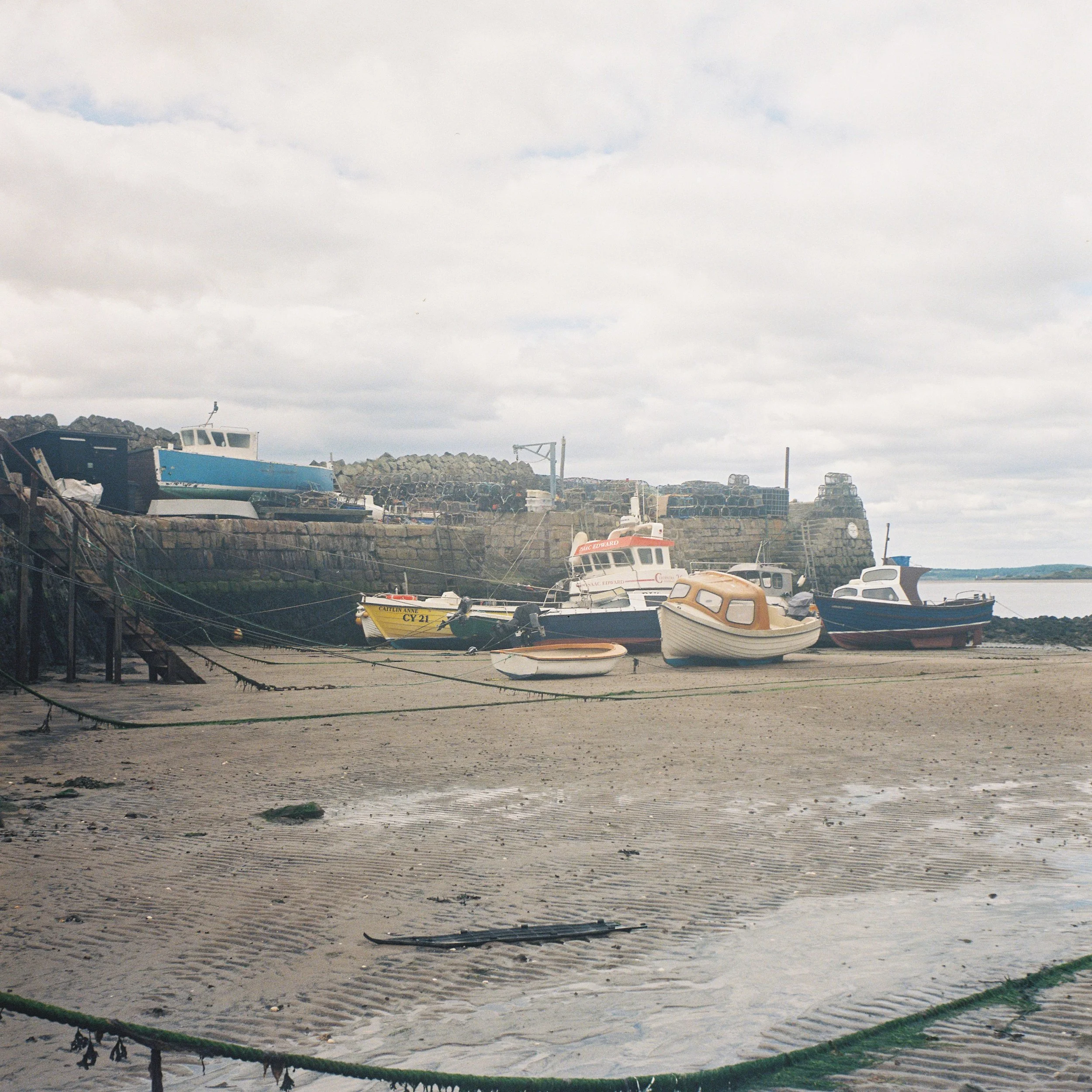 Boats docked on a sandy beach under cloudy sky, with some structures and rocks in the background.