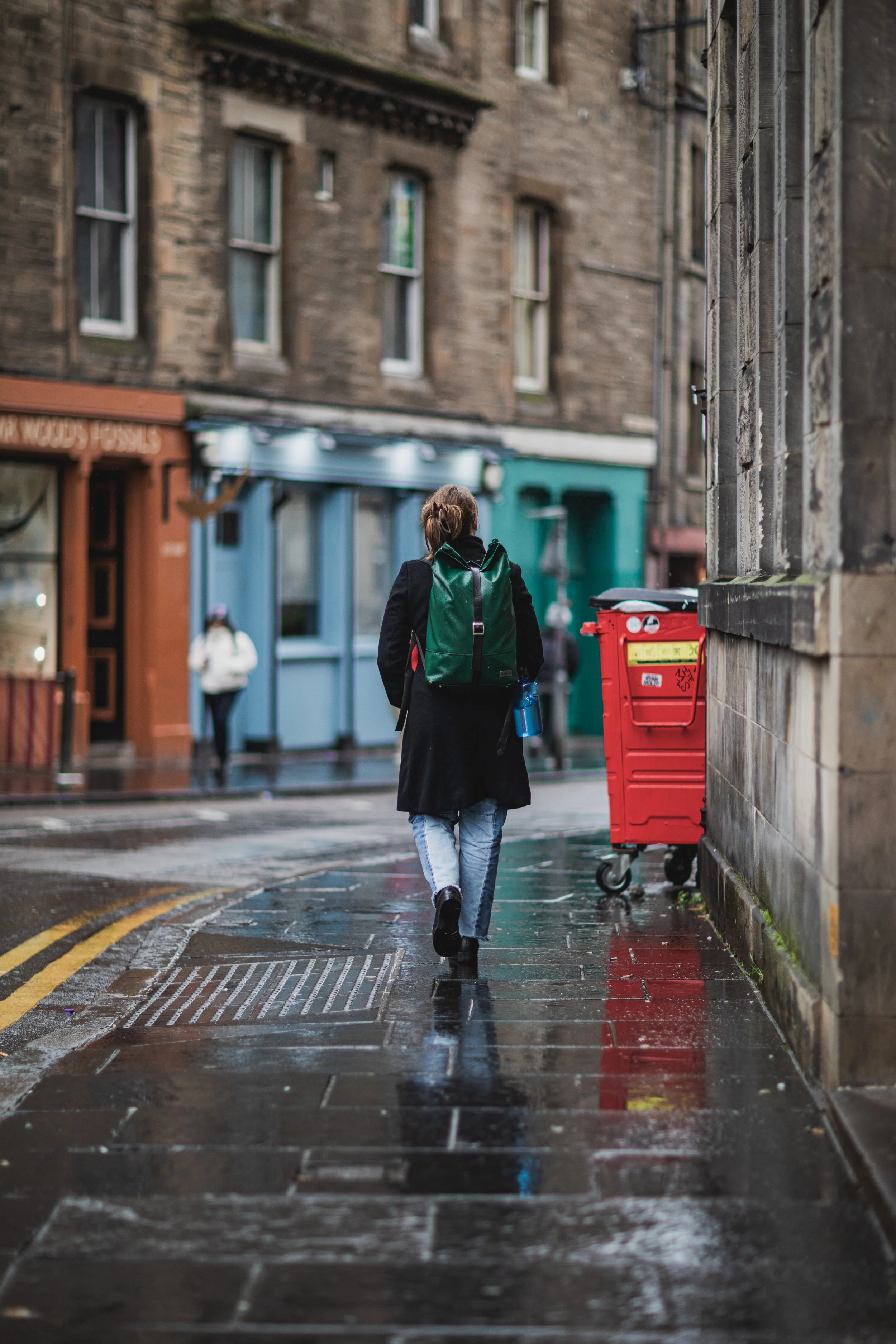 A person walking on a wet city sidewalk with a green backpack, wearing a long black coat and light blue jeans, near a red trash bin, on a rainy evening.