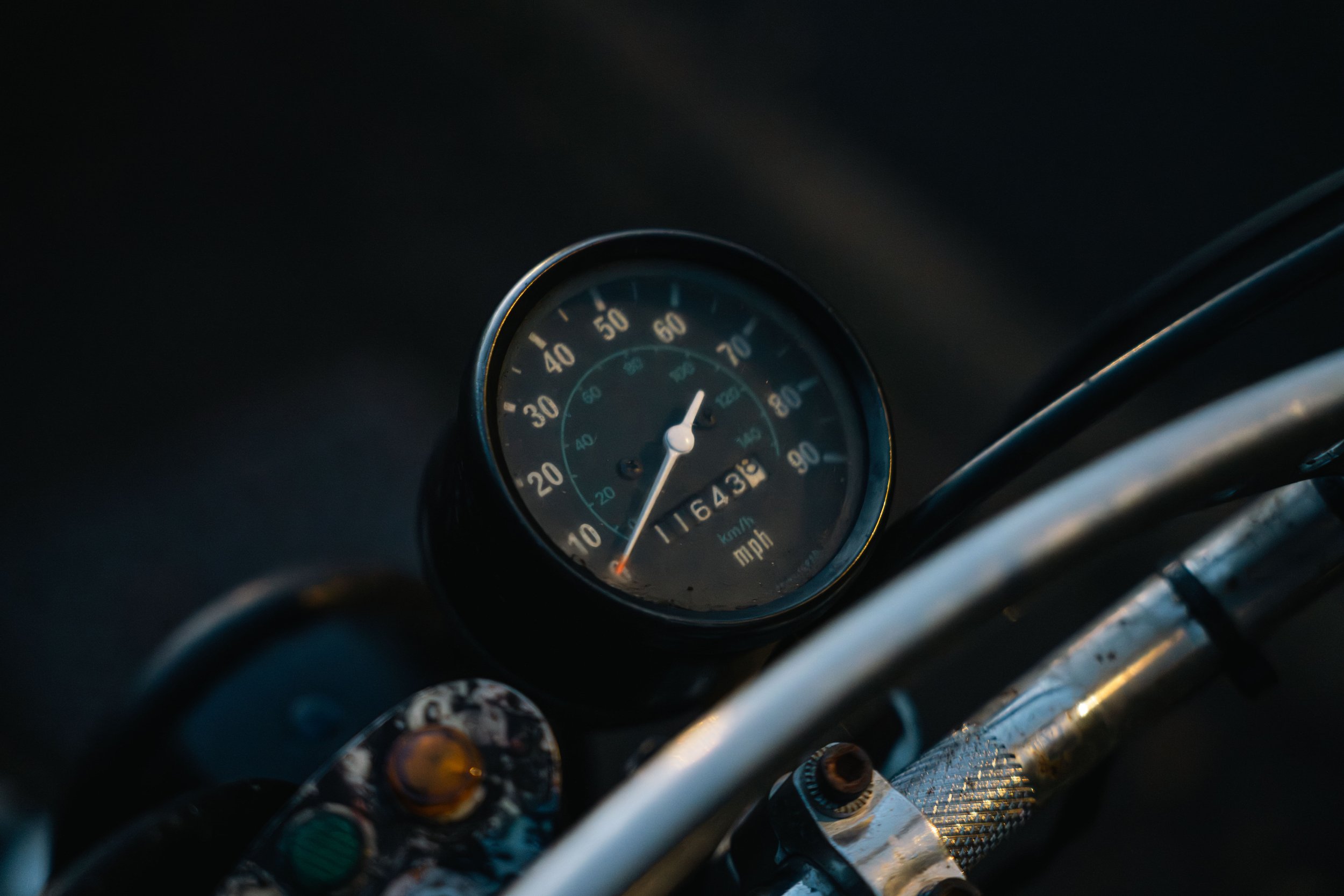 Close-up of a vintage car speedometer indicating a speed of approximately 65 mph, with various other car parts visible in the background.