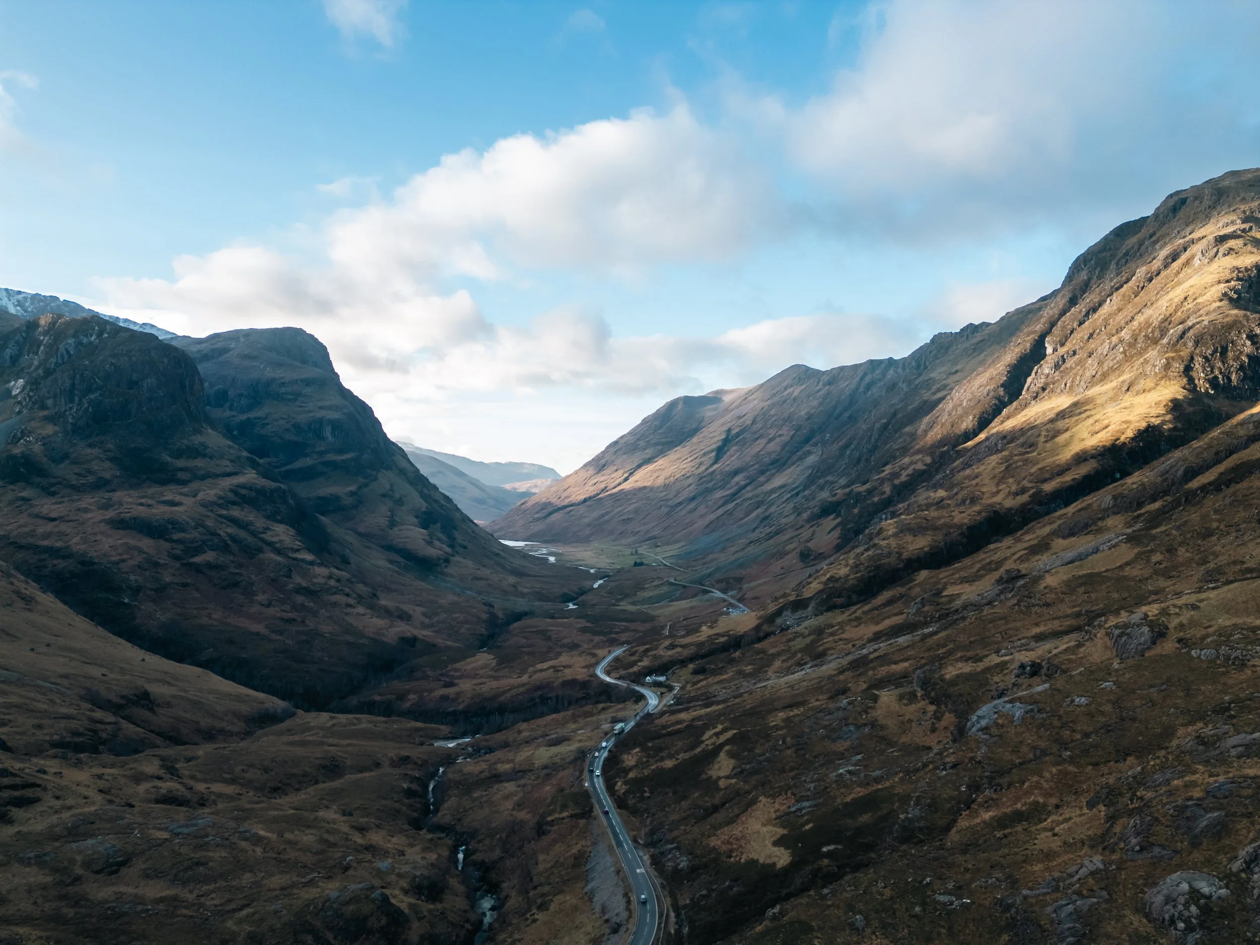 A scenic view of snow-capped mountains on the left and rugged hills on the right, with a narrow winding road running through a valley with a river flowing below and beneath a partly cloudy sky.
