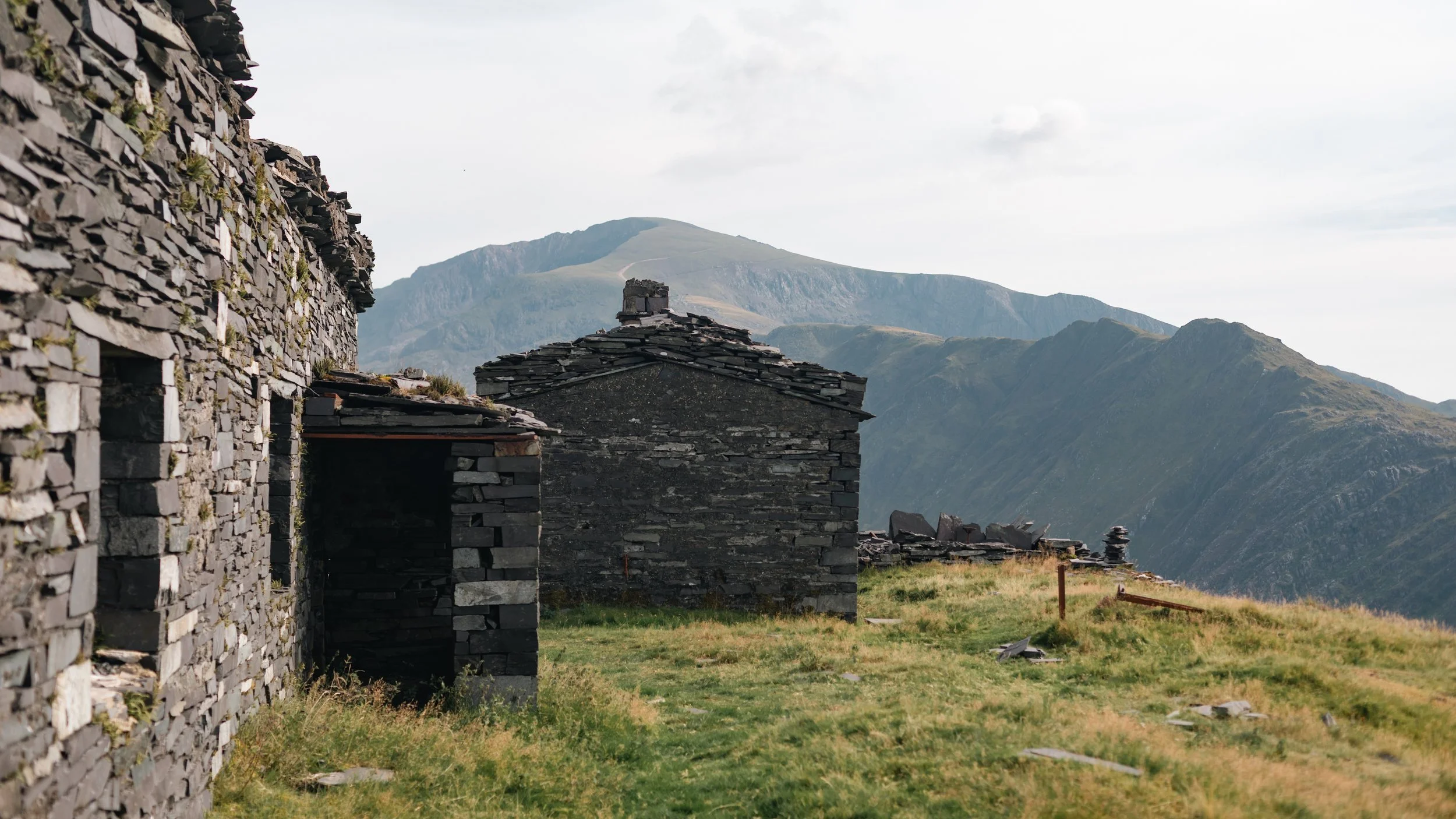 Old stone buildings with slate roofs in a mountainous landscape with green hills and a cloudy sky.