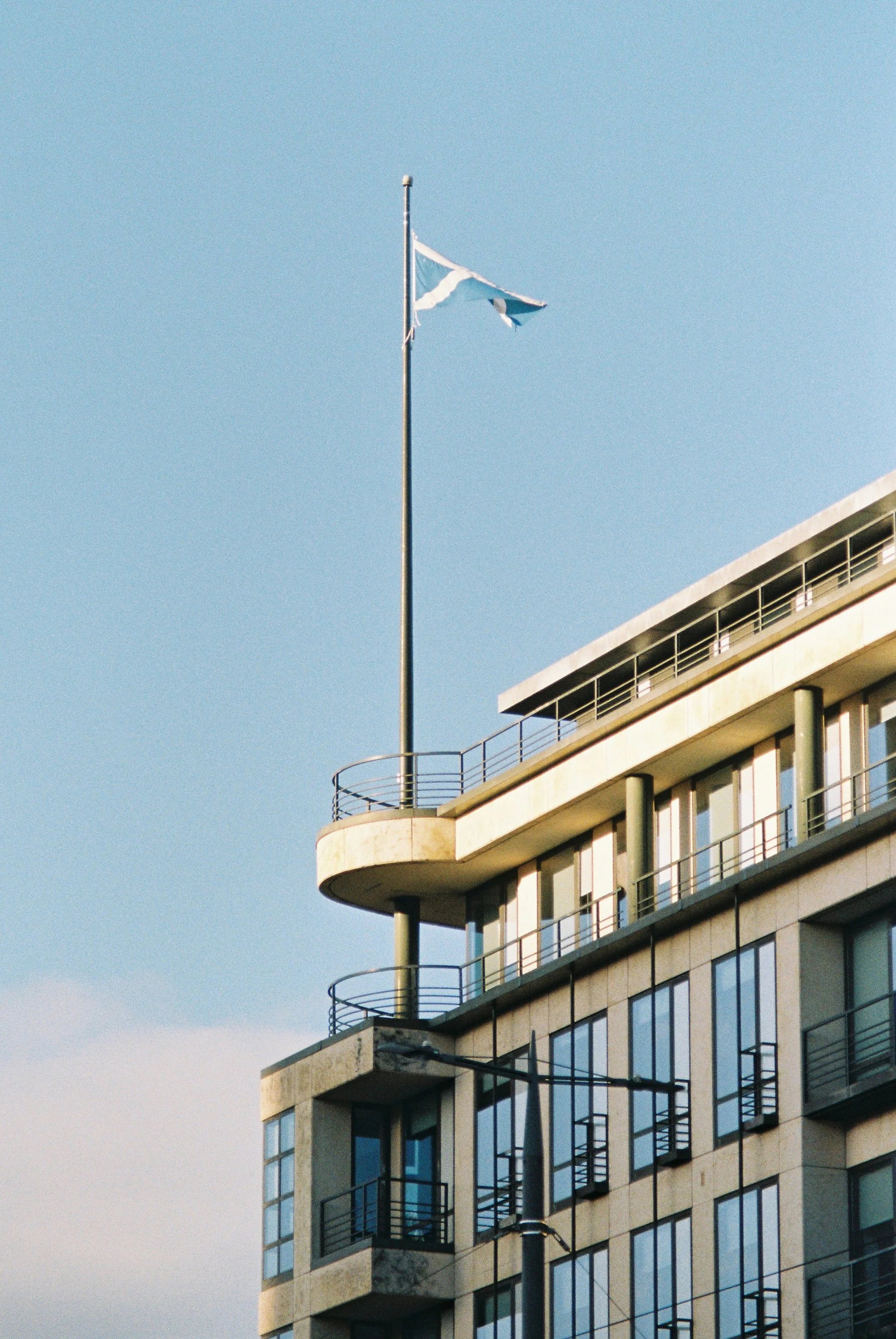 A high-rise building with glass windows and balconies under a clear sky, with a flagpole and tattered flag on top.
