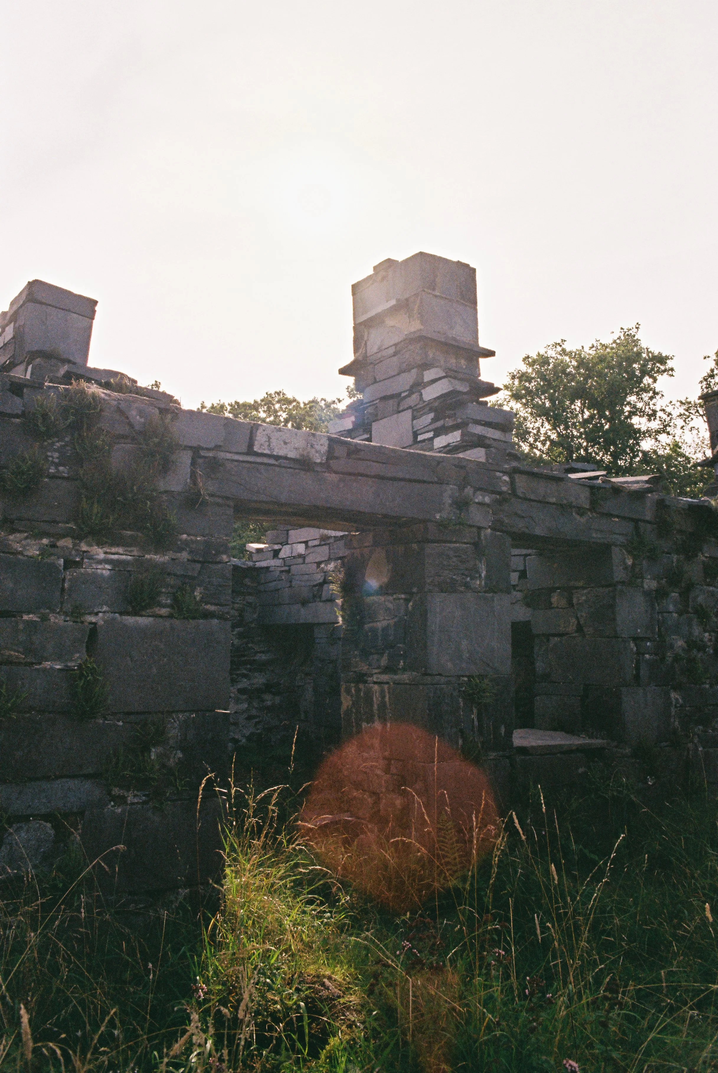 Ruined stone chimney and structures in a grassy, overgrown area, with trees in the background and the sun shining brightly overhead.