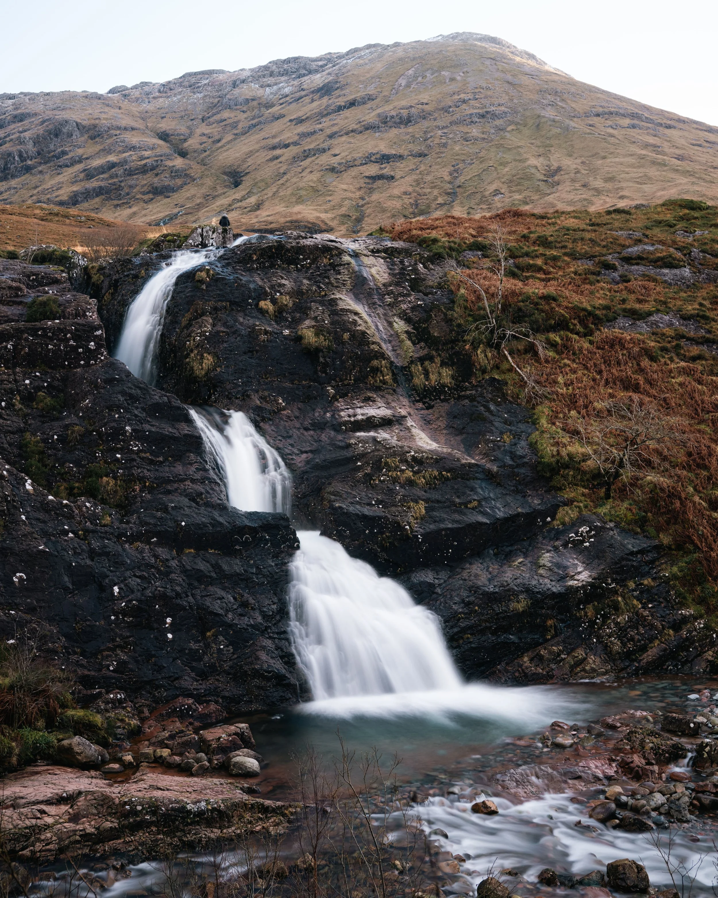 A scenic landscape featuring a mountain in the background, a waterfall cascading over rocks in the middle, and a rocky stream in the foreground.