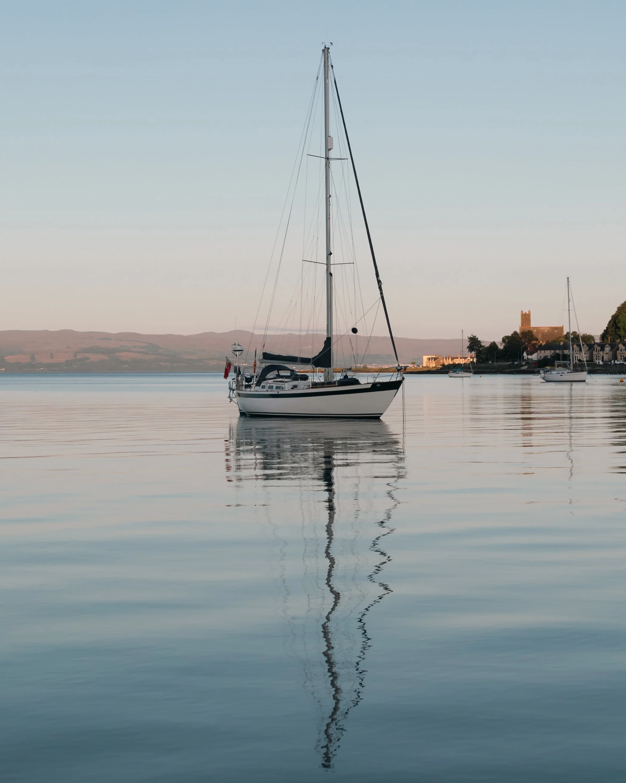 A sailboat floating on calm water with reflections. In the background, a historic building and other boats are visible along the shoreline, with hills and a clear sky.