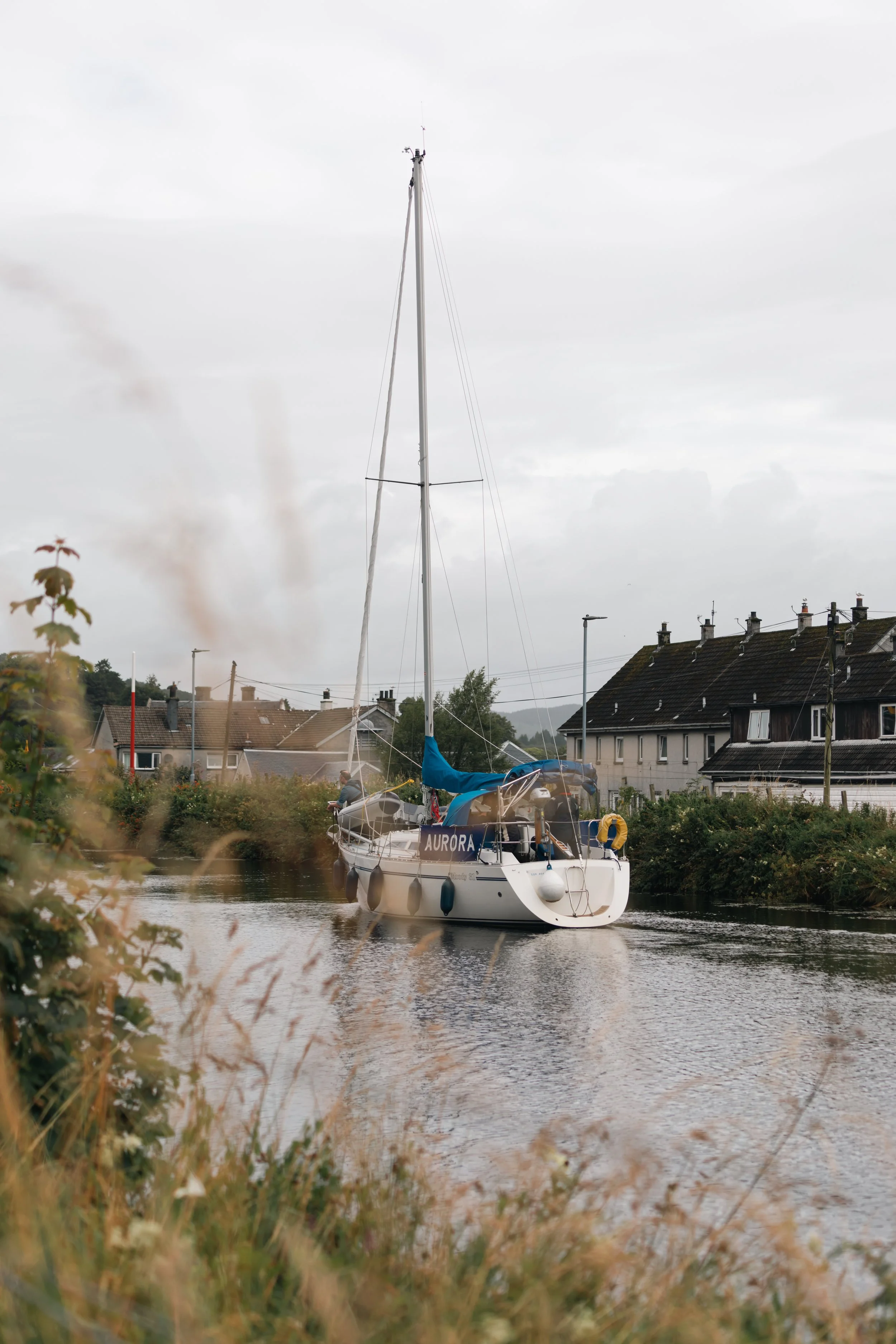 A sailboat named Aurora is on a calm waterway near houses with dark roofs and chimney stacks, under a cloudy sky.