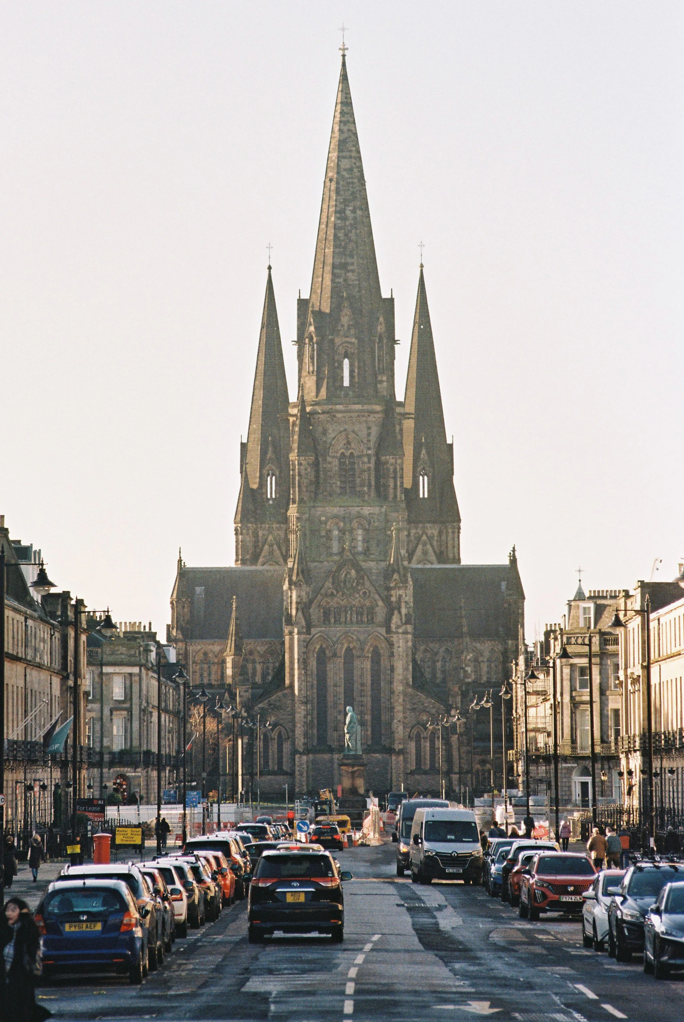 Street view in front of a large gothic church with a tall spire, surrounded by cars and people, with buildings on either side.