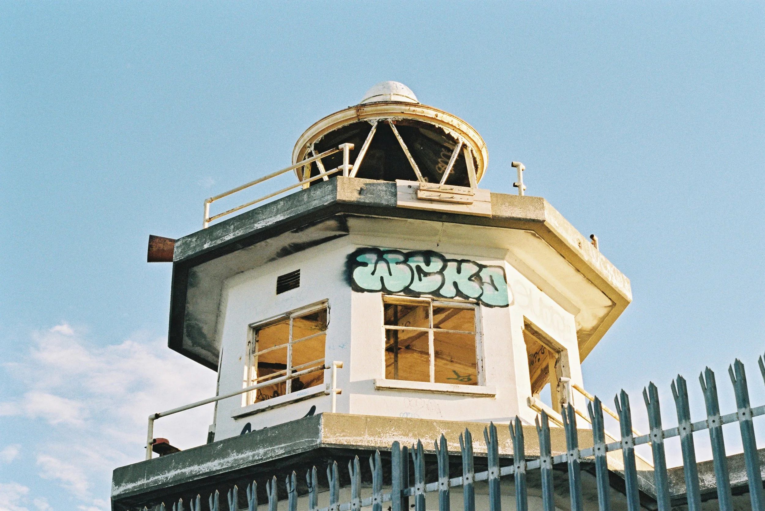 An old, weathered lighthouse with chipped paint and graffiti on its walls, surrounded by a spiked metal fence, under a clear blue sky.