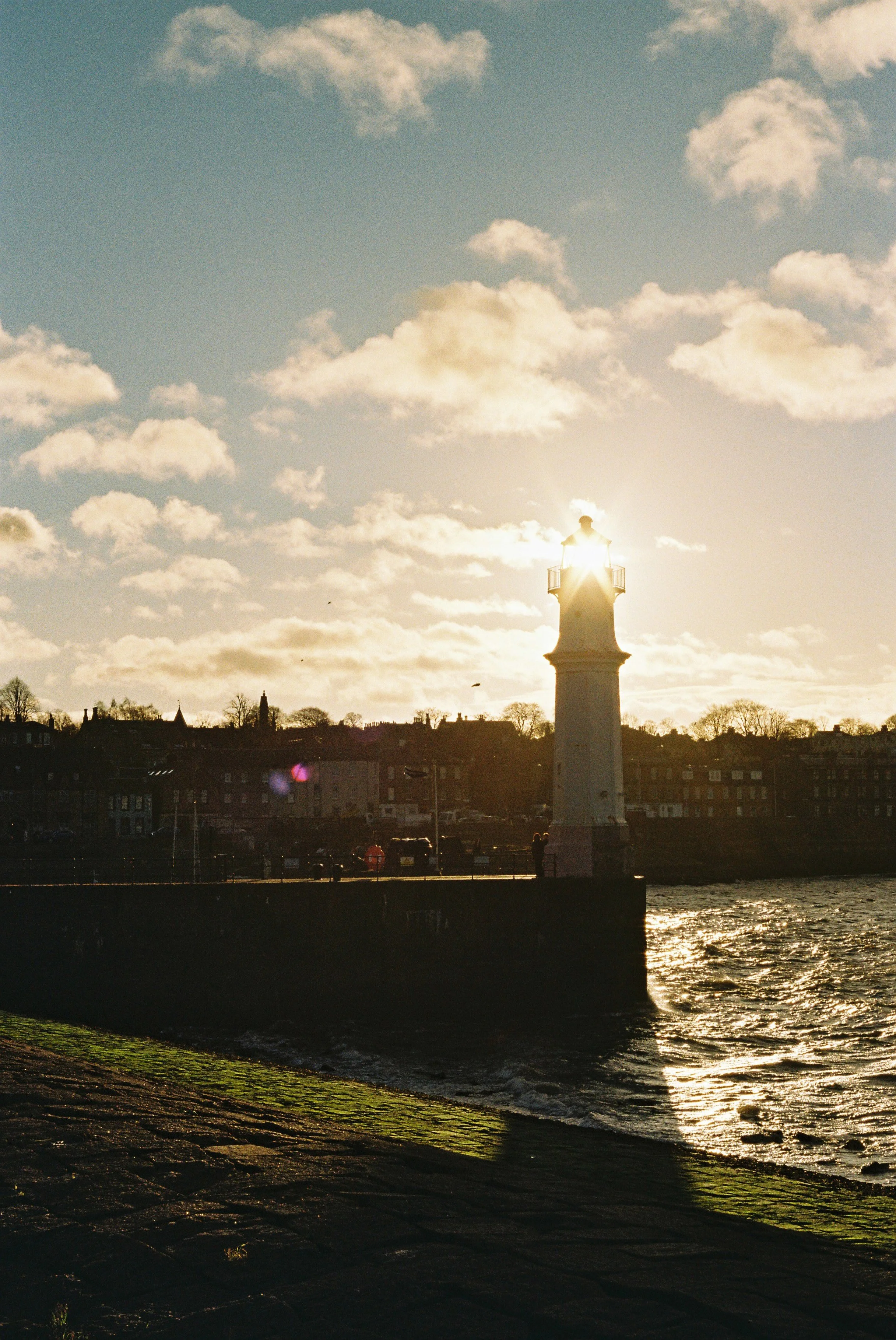 A lighthouse with the sun behind it, casting a glow in the sky filled with clouds, and water in the foreground reflecting the light.