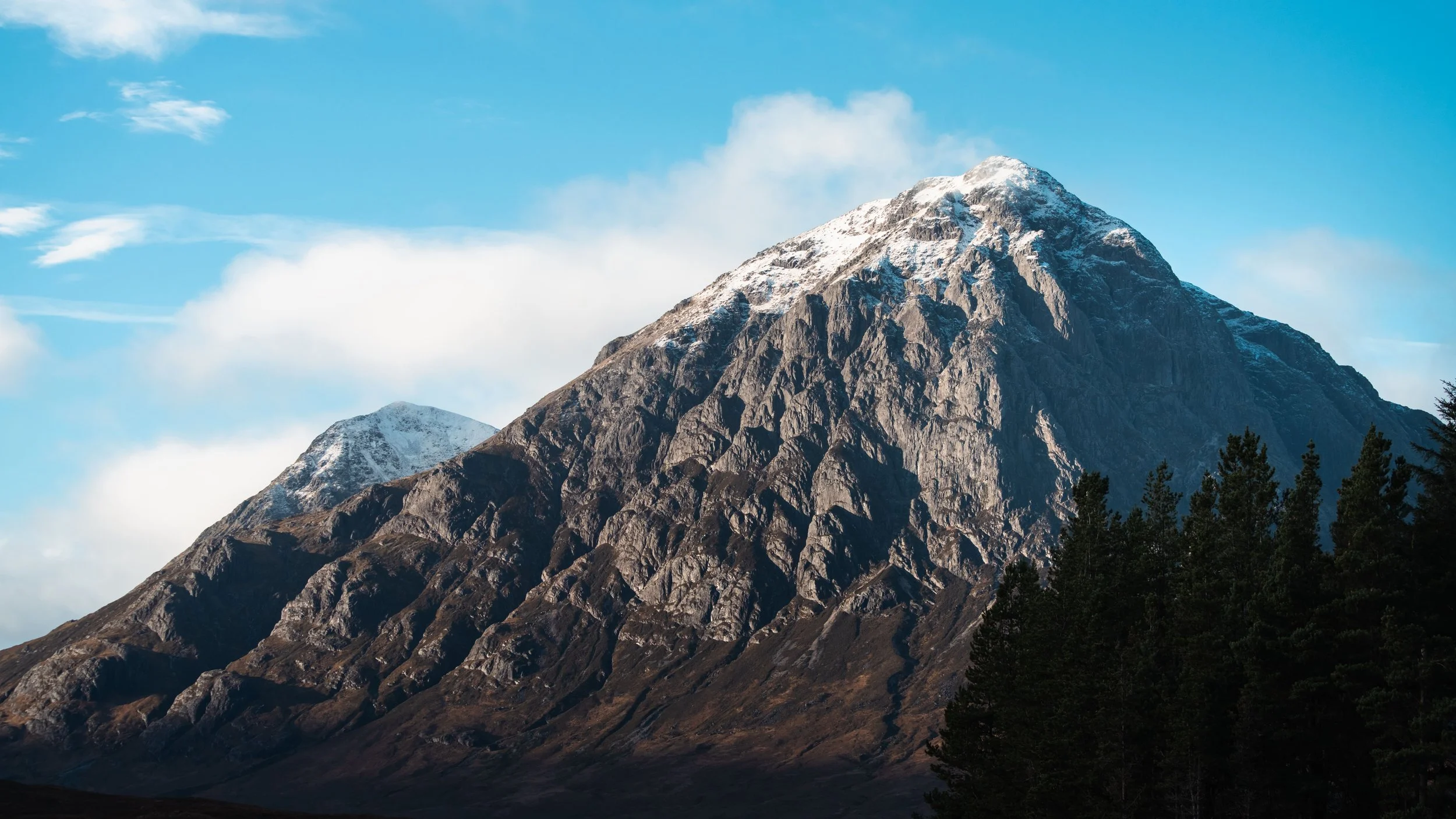 A mountain with snow on the peak and rugged, rocky slopes surrounded by a few green trees at the base, under a blue sky with some clouds.