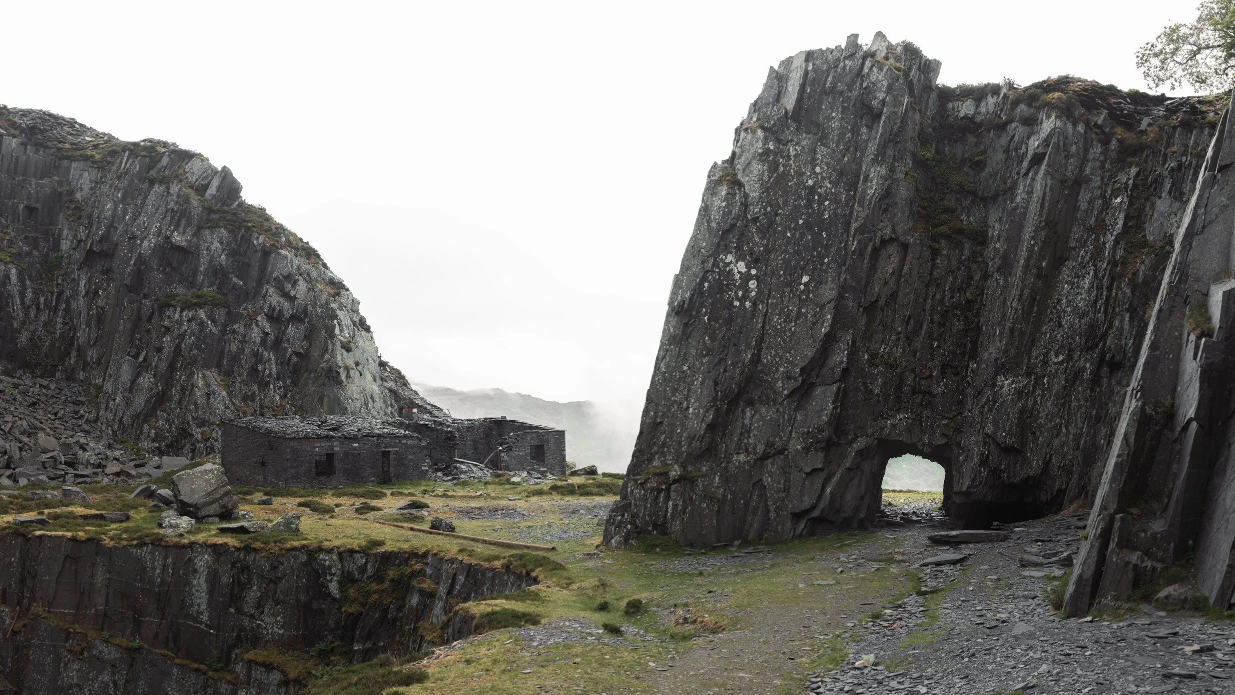 A scenic view of tall rocky cliffs with a small stone building or house at the bottom, and a natural arch formation in one of the cliffs, under a cloudy sky.